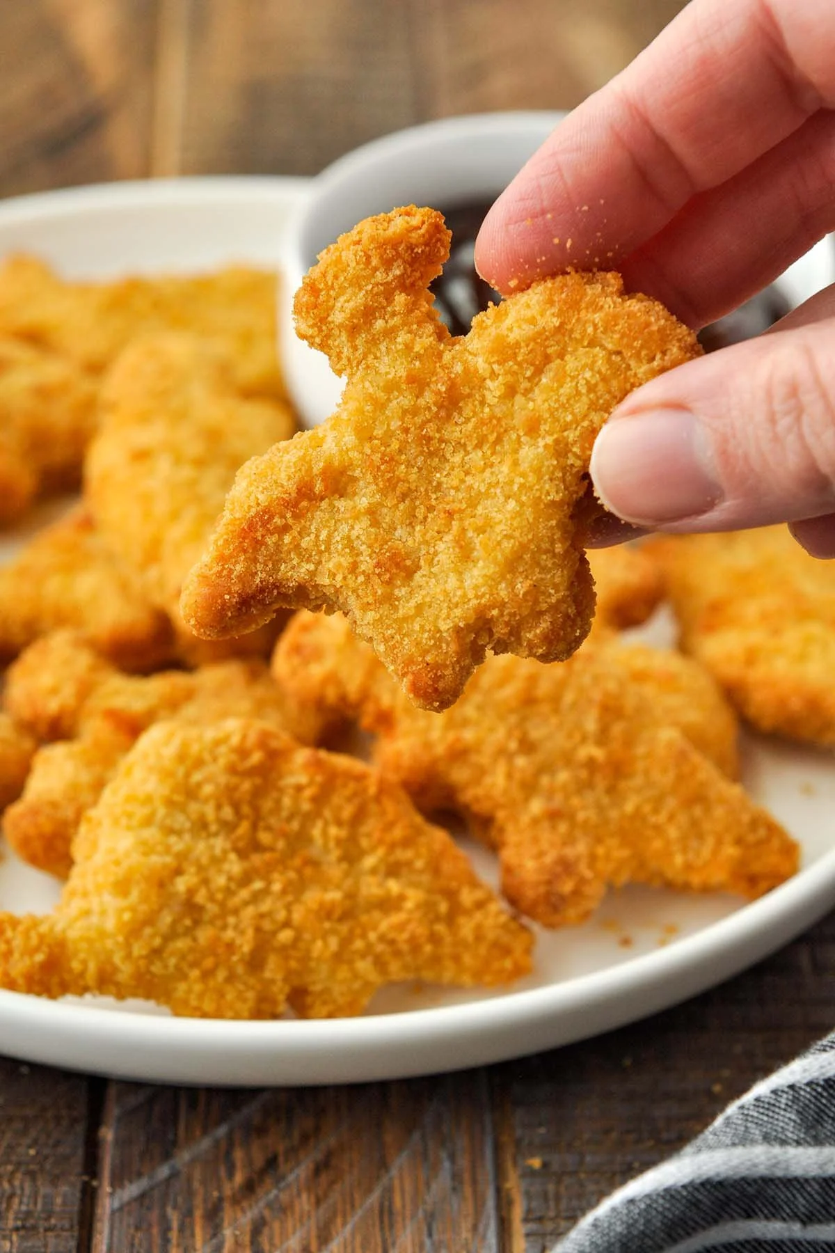 Fried chicken nugget held between thumb and forefinger above a bowl of similar chicken nuggets on a wooden table.