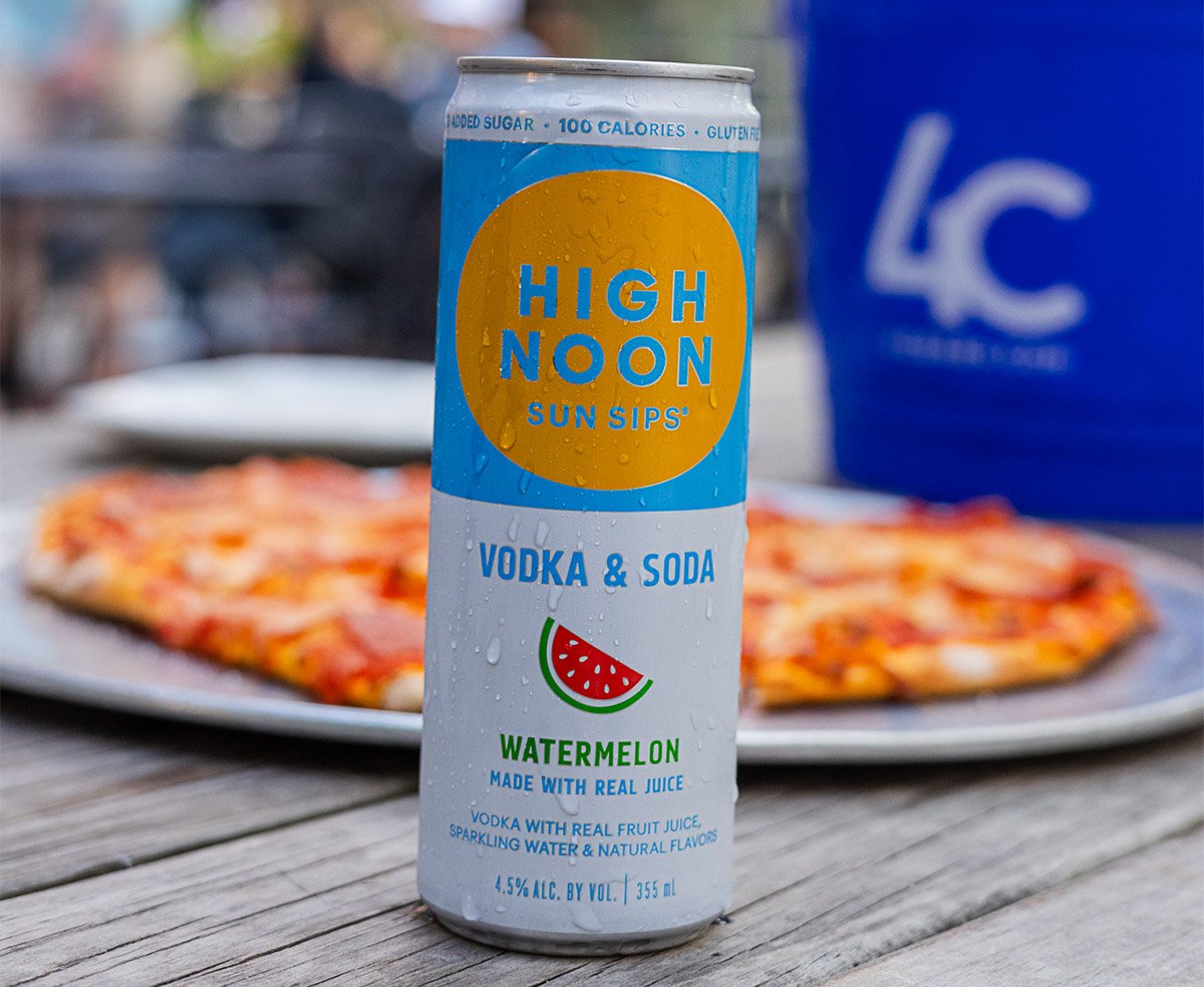 A can of High Noon Vodka & Soda Sunset Sips Watermelon flavored vodka and soda with real juice, placed on a wooden table outdoors, with a pizza in the background.