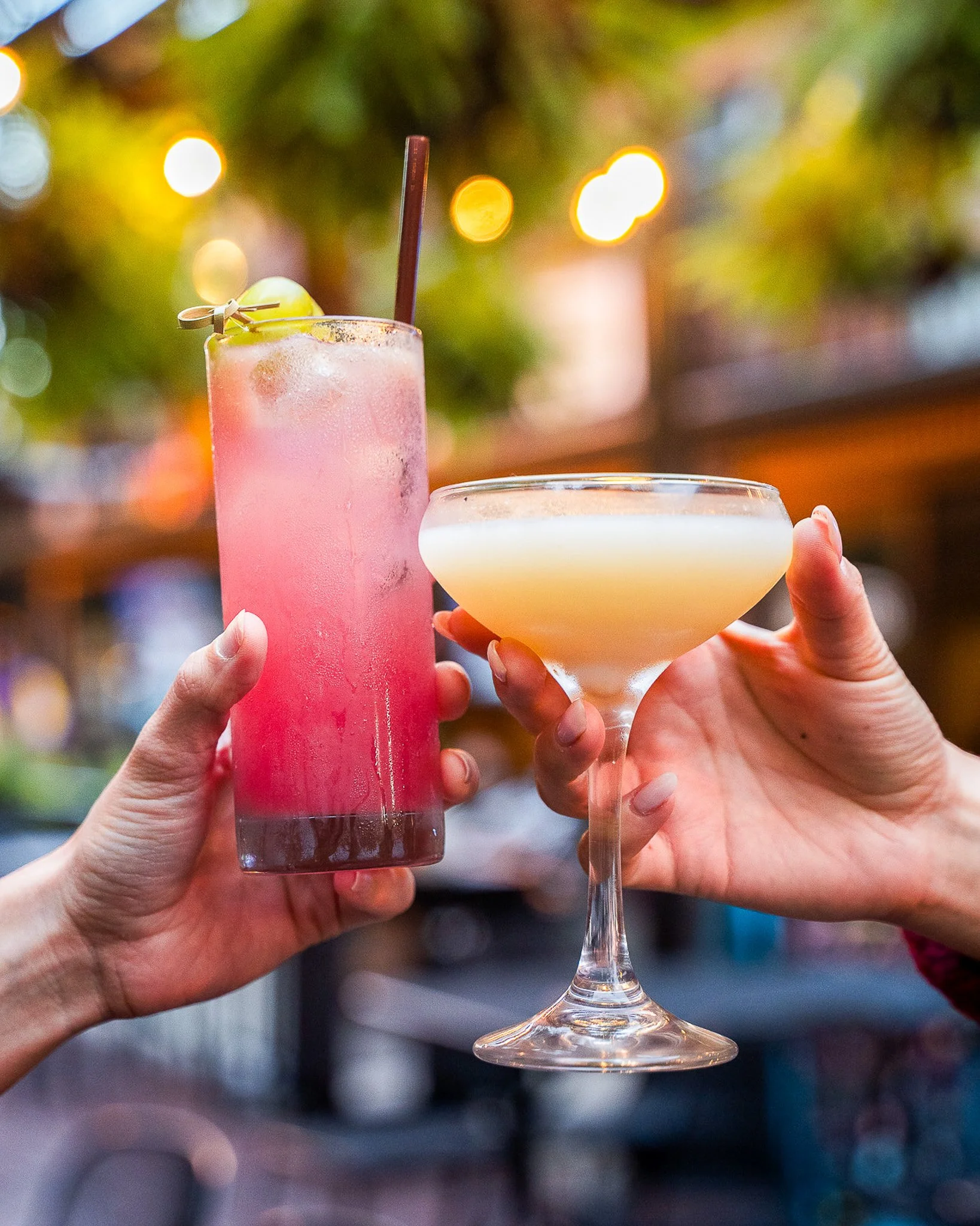 Close-up of a person holding a pink cocktail with a lime wedge and a straw, and another person holding a cream-colored cocktail in a coupe glass, with a blurred background of colorful lights and greenery.