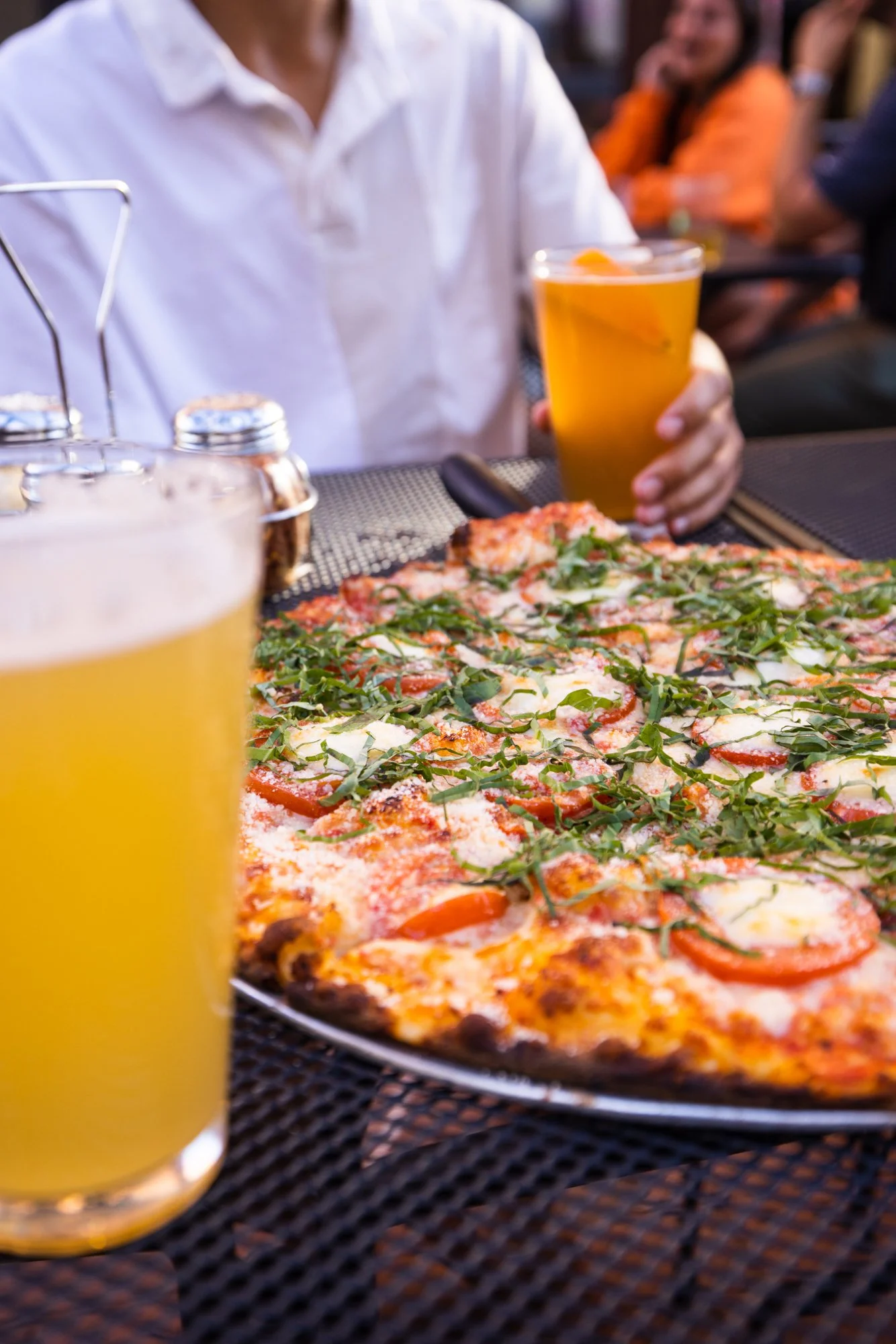 A person sitting at an outdoor table holding a glass of beer, with a pizza topped with tomatoes, cheese, and green herbs in front of them, and other people in the background.