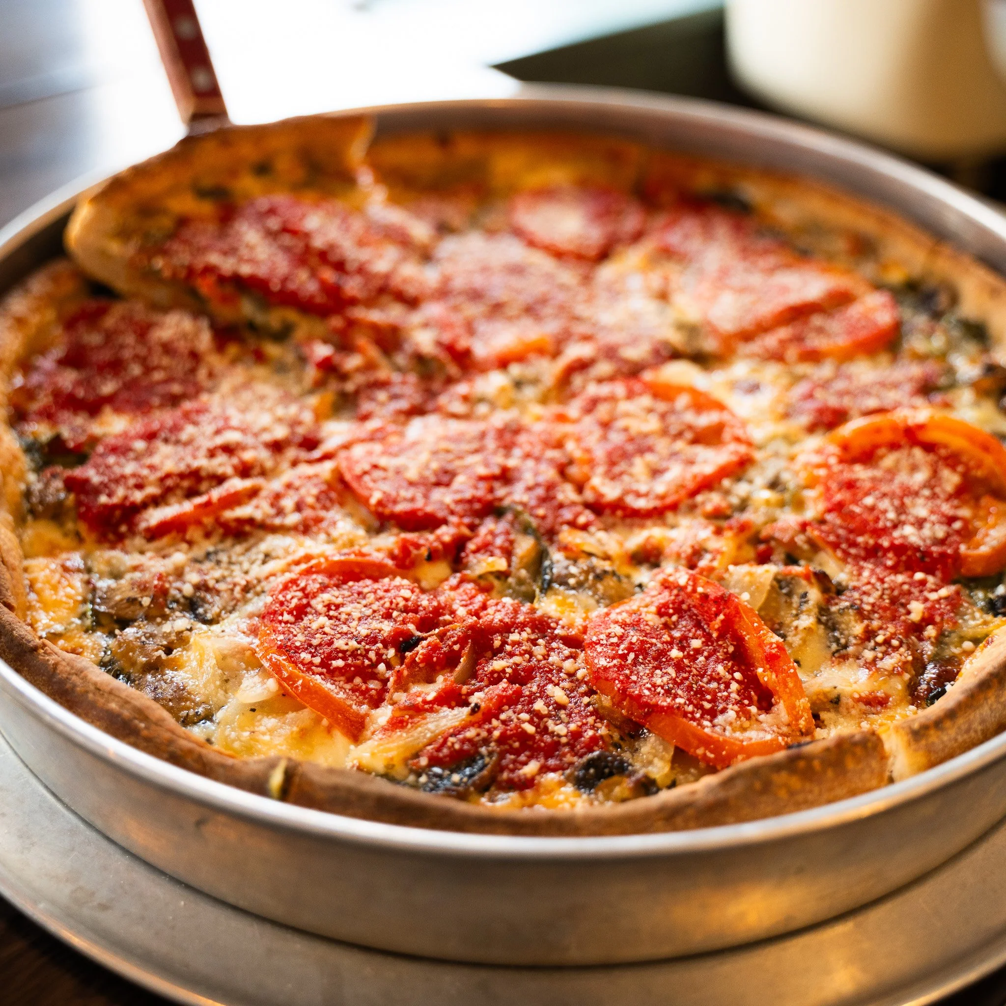 Close-up view of a pizza in a metallic oven or air fryer, showing toppings including pepperoni, cheese, and vegetables, with a slightly blurred background.