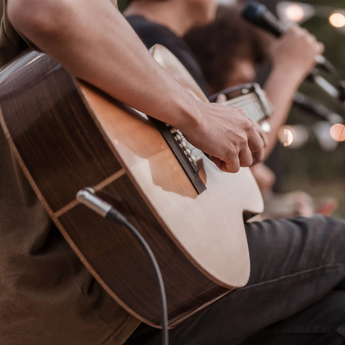 Close-up of a person playing an acoustic guitar, with focus on their hand strumming and the guitar's body, and another person in the background also holding a guitar.