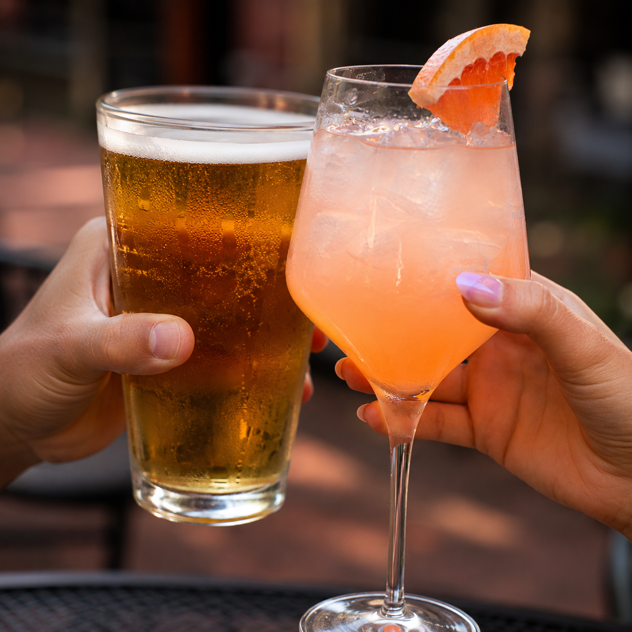 Two hands holding drinks: a pint glass of beer and a pink cocktail with a grapefruit garnish, clinking together.