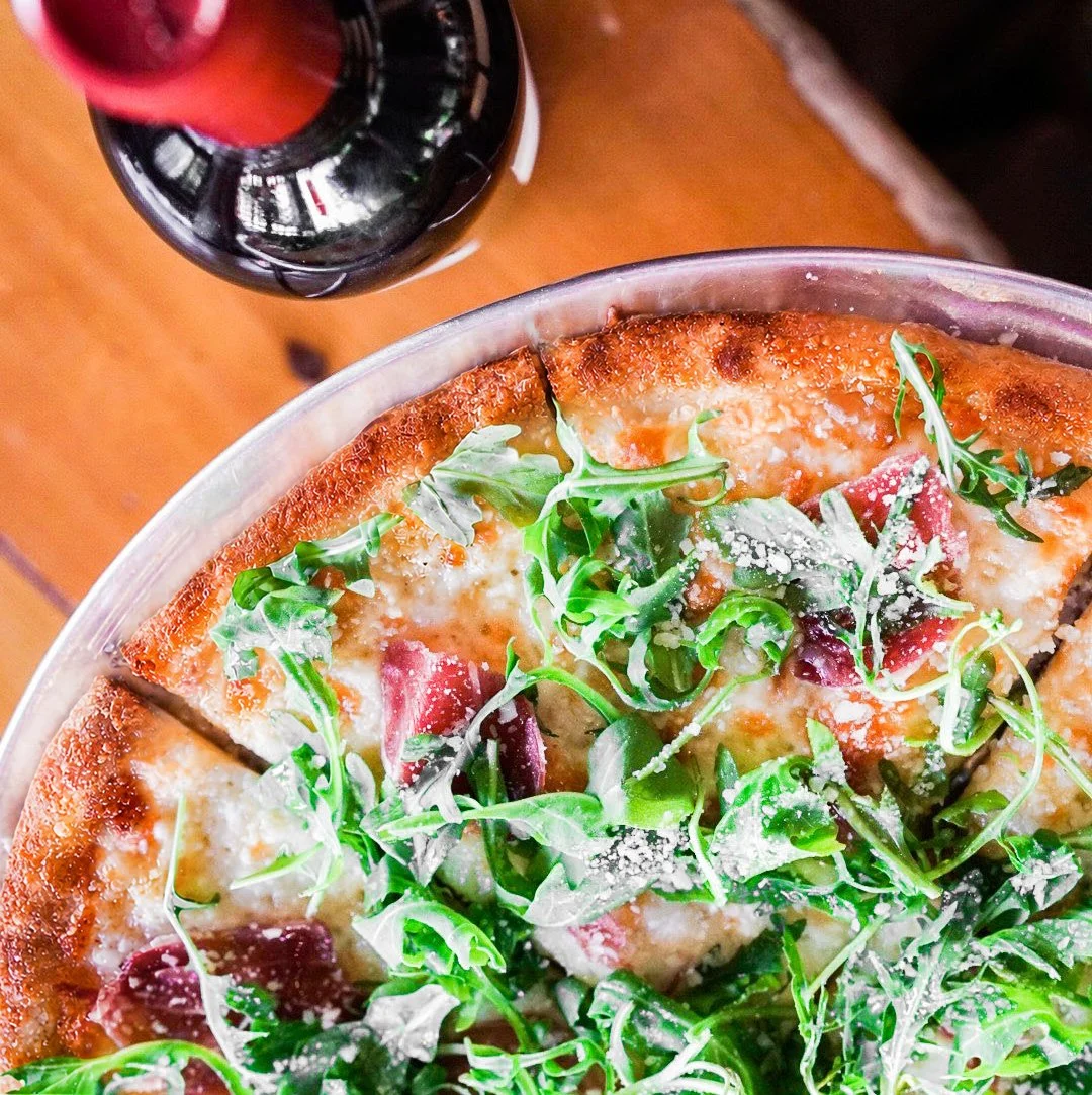 A pizza with arugula, grated cheese, and slices of meat, topped with a splash of grated cheese, next to a glass of dark soda, on a wooden table.