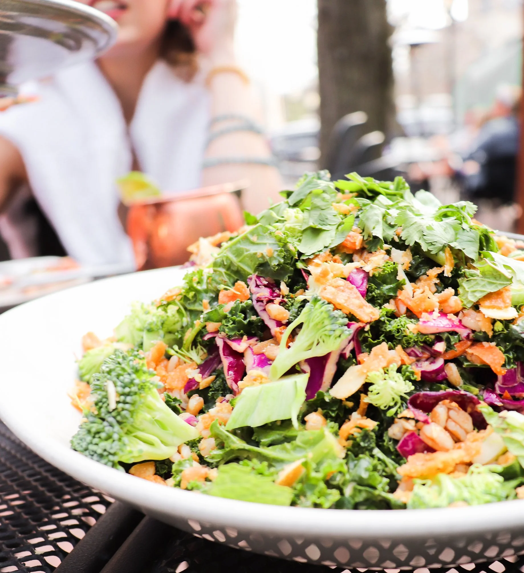 A colorful salad with mixed greens, shredded vegetables, and toppings, served in a white bowl outdoors at a restaurant or picnic.