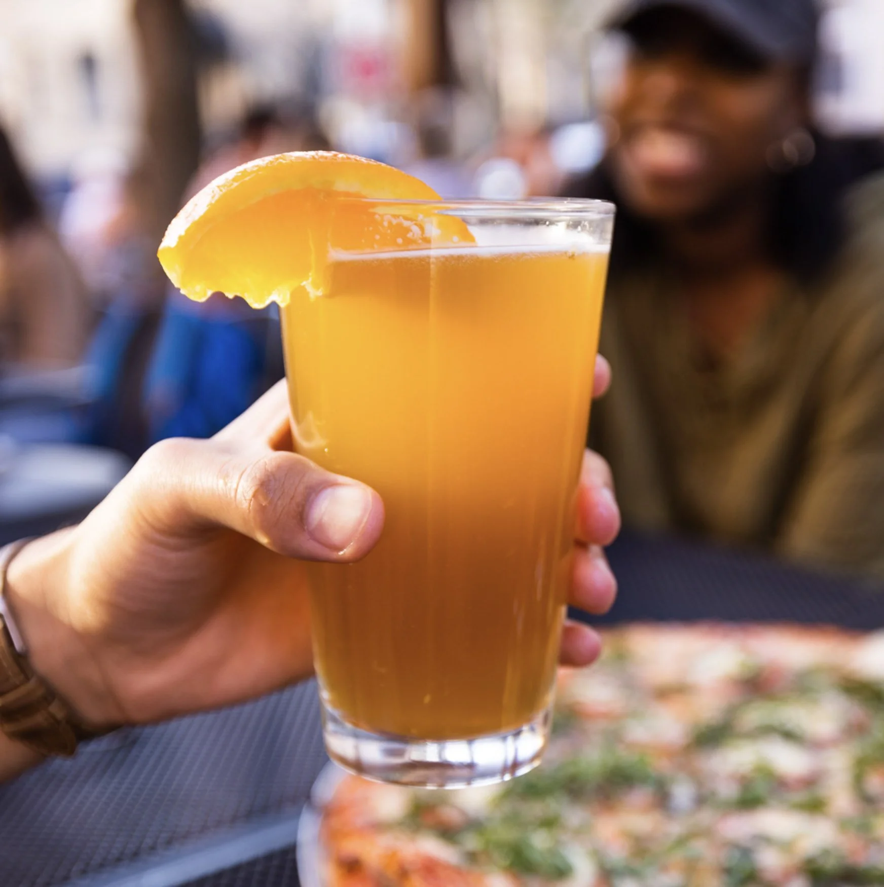 A clear glass of orange juice with an orange wedge garnish, being held by a person outdoors during daytime, with blurred people and autumn leaves in the background.