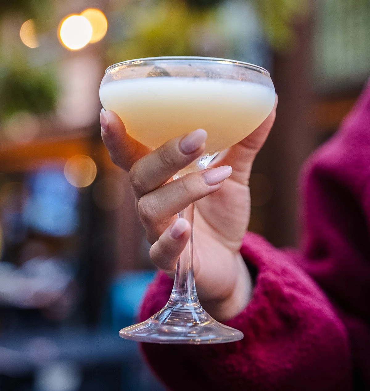 A person holding a coupe glass filled with a pale yellow cocktail, with blurred colorful lights in the background.