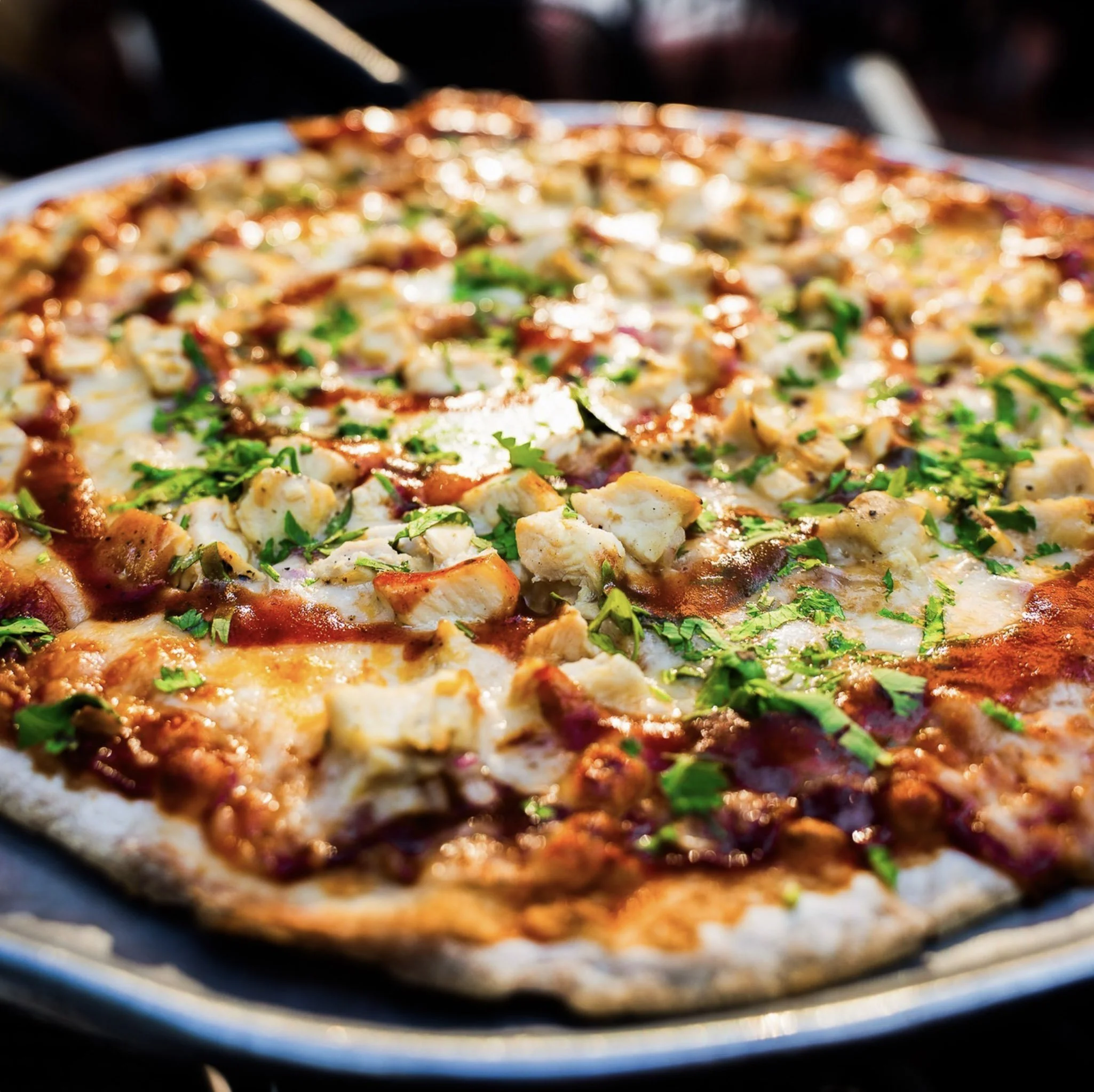 Close-up of a pizza with toppings like chicken and herbs, viewed from a side angle in a blurry background.