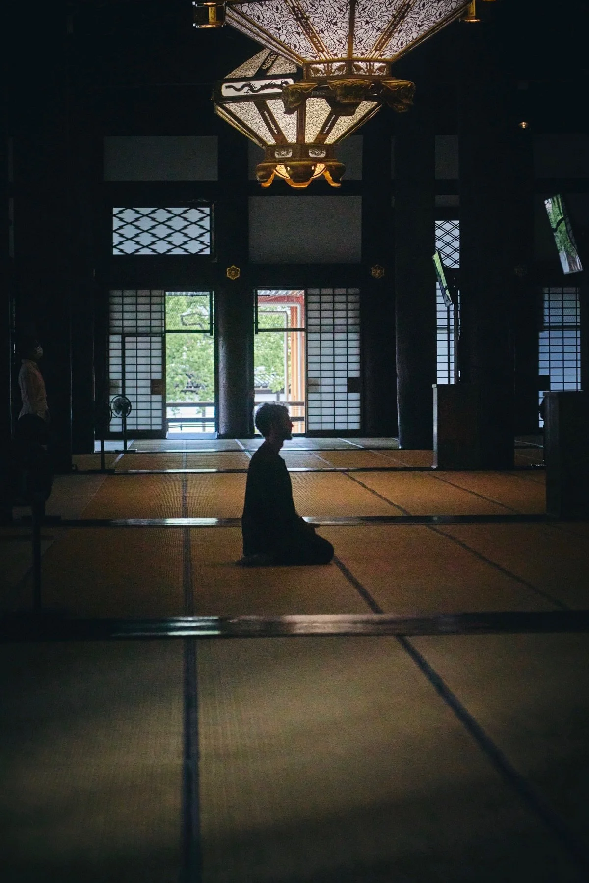 Inside a traditional Japanese room with tatami mats, a person is kneeling in meditation with lamps and shoji screens in the background, illuminated by natural light from open sliding doors.