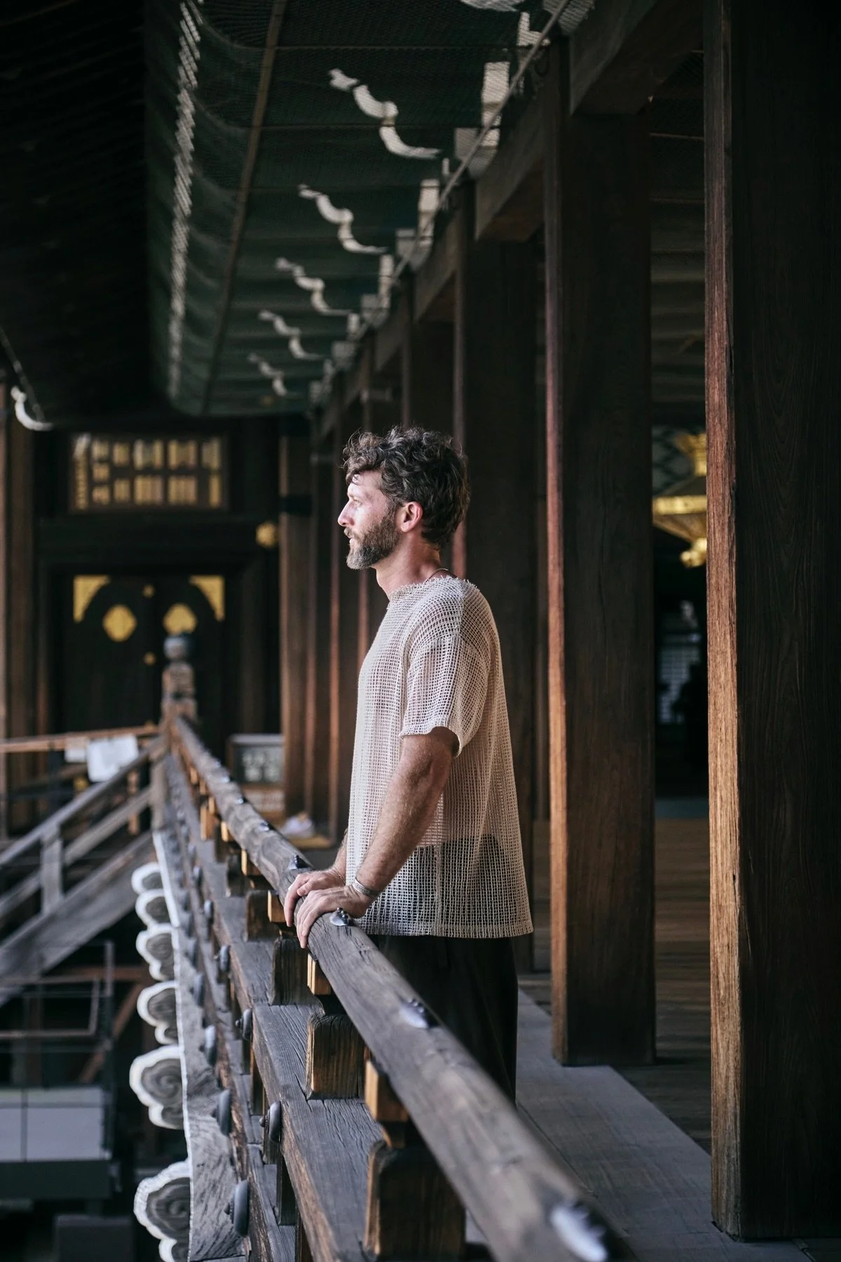 A man with curly hair and a beard stands on a wooden balcony, looking outward. He is wearing a beige, mesh short-sleeved shirt and black pants, with his hands resting on the railing. The background features wooden pillars and a dark ceiling with decorative elements.