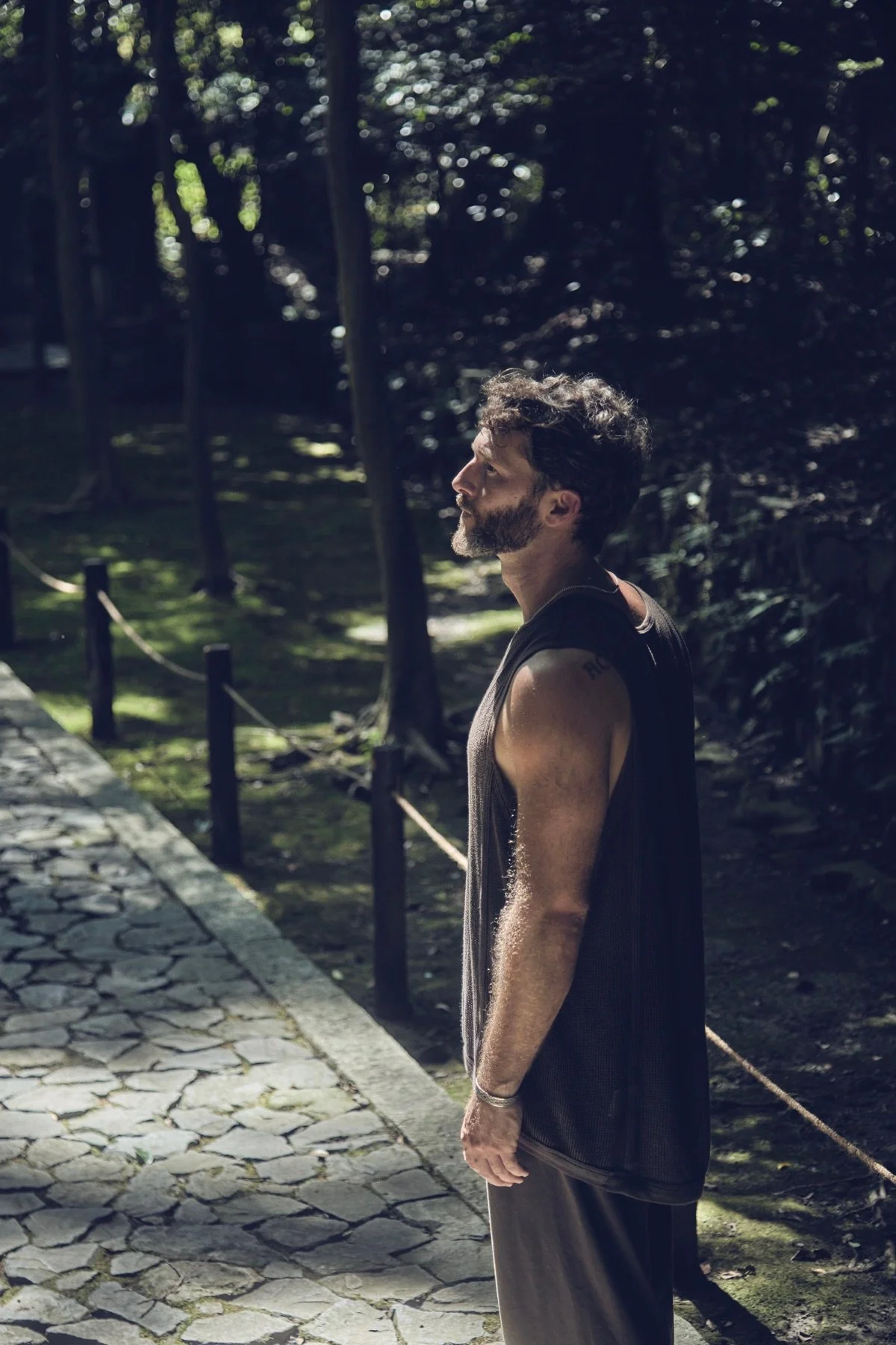 A man with dark, curly hair and a beard, wearing a black sleeveless shirt and gray pants, stands outdoors on a stone pathway with trees and sunlight filtering through the foliage in the background.