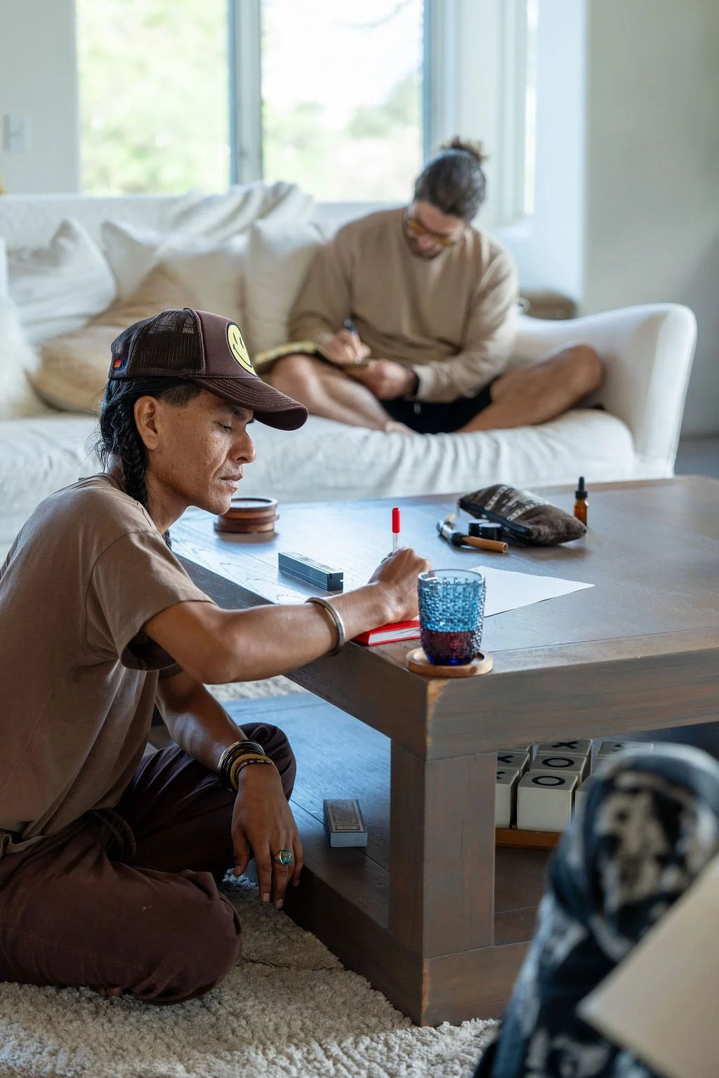 Two men in a living room, one sitting on the floor and one on the couch, surrounded by items such as a notebook, writing tools, and a glass of water.