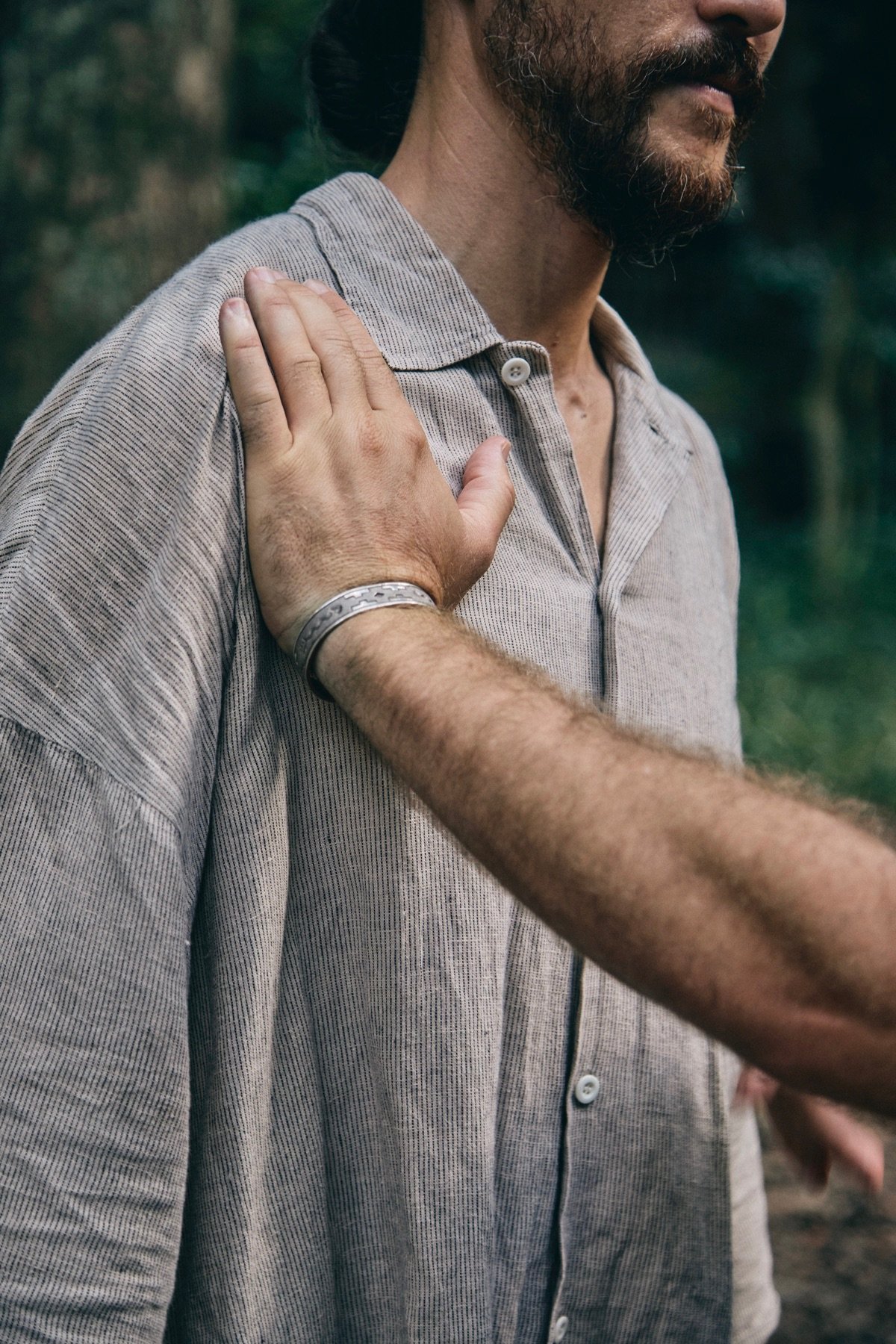 A man with a beard and mustache wearing a beige striped button-up shirt with a silver bracelet on his wrist, outside in a natural setting, placing his right hand on his shoulder.