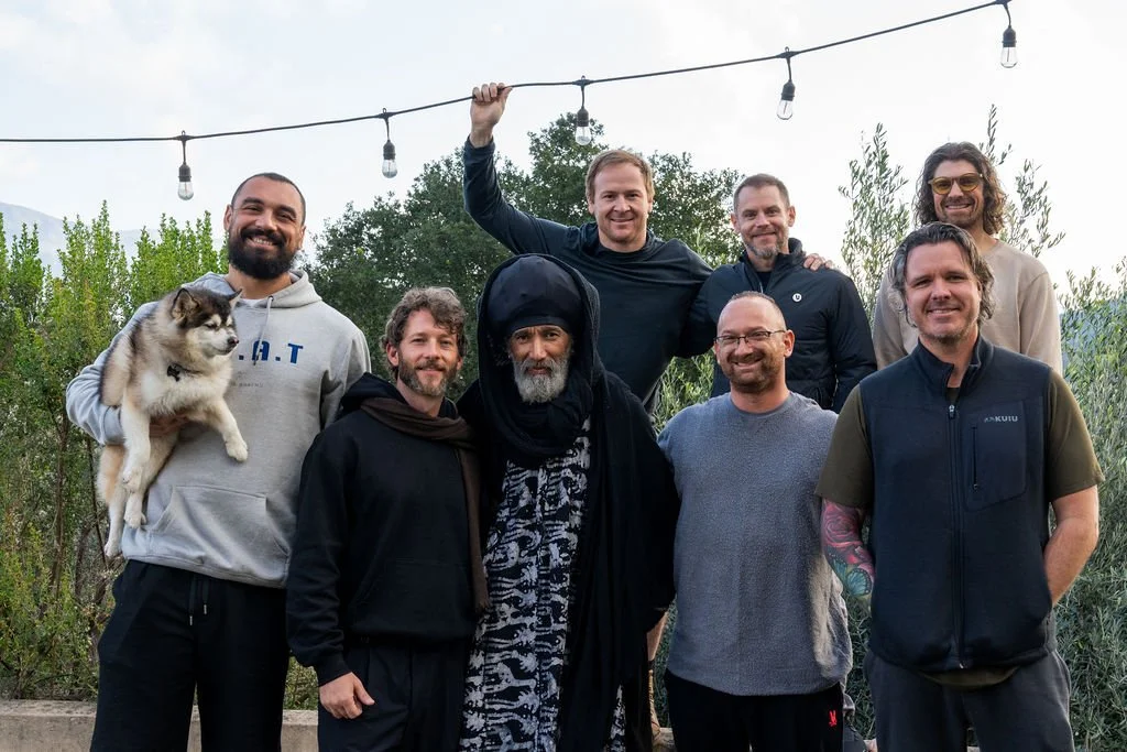 A group of nine men, one holding a dog, standing outdoors. Some are smiling, with string lights overhead and trees in the background.