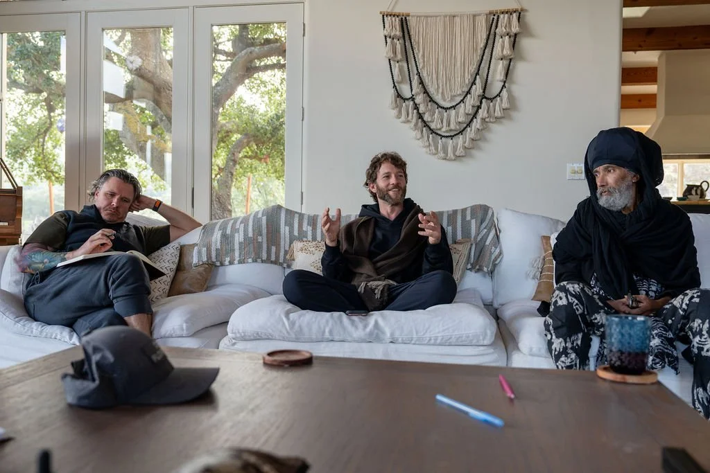Three men sitting on a white couch in a bright living room with large windows and green trees outside. The man on the left is leaning back and writing in a notebook, the man in the middle is talking and gesturing, and the man on the right is wearing a headscarf and dark clothing. There are pens and a hat on the wooden table in front of them.