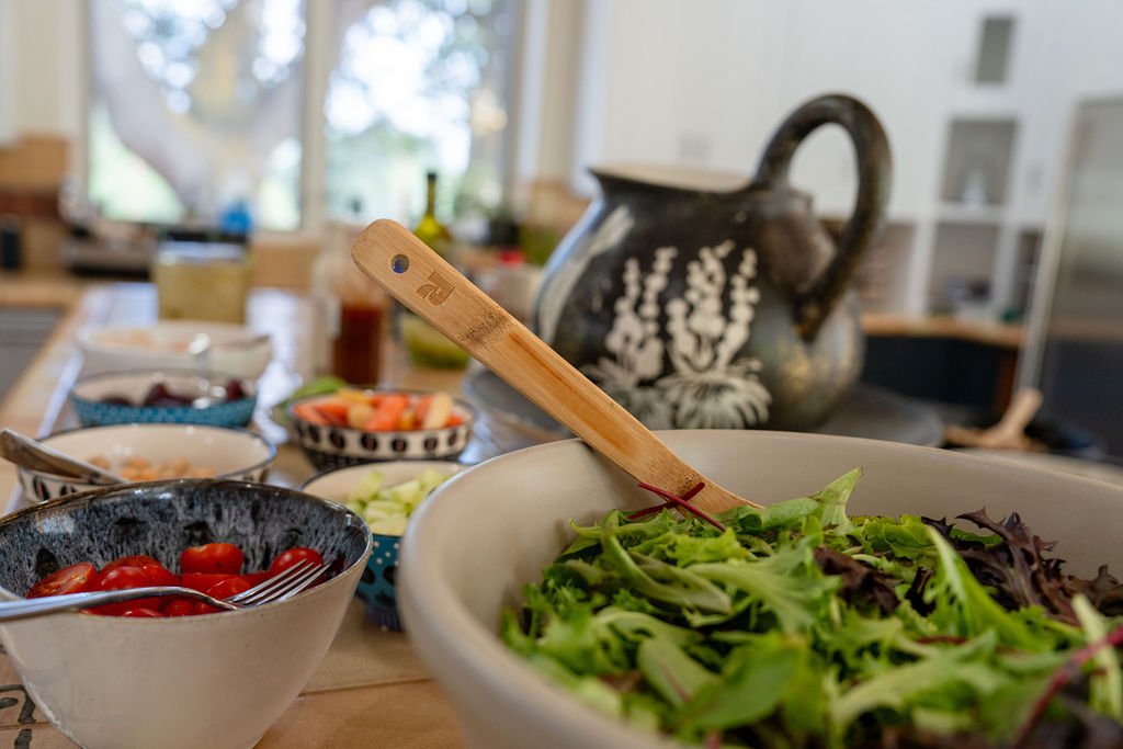 Fresh salad in a white bowl on a dining table with various bowls of vegetables and toppings, a ceramic teapot in the background.