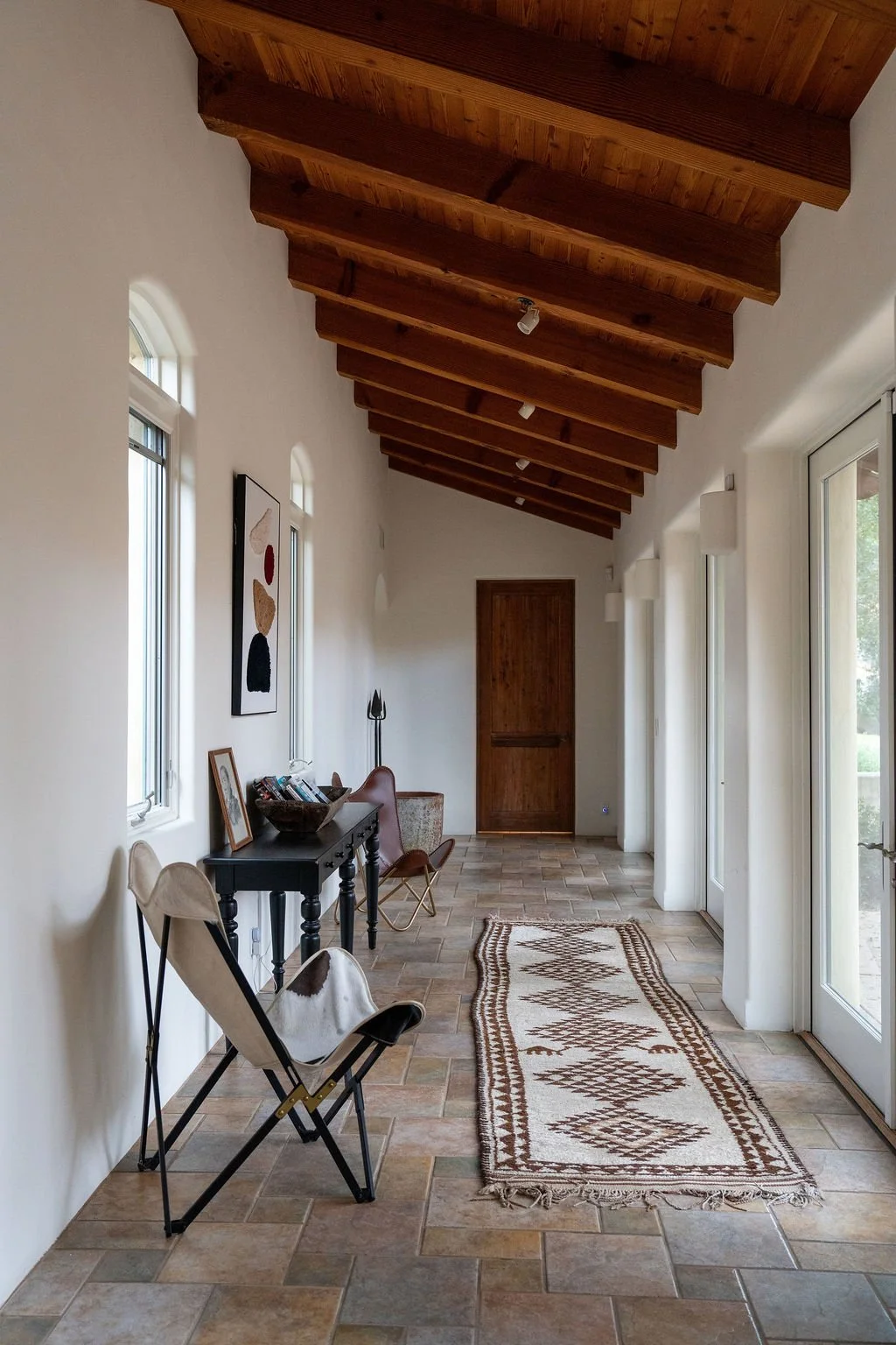 A bright, narrow hallway with a wooden vaulted ceiling, white walls, tiled floor, and a patterned rug. Contains chairs, a black console table with framed photos, and artwork on the wall. Large glass door on the right allows natural light.