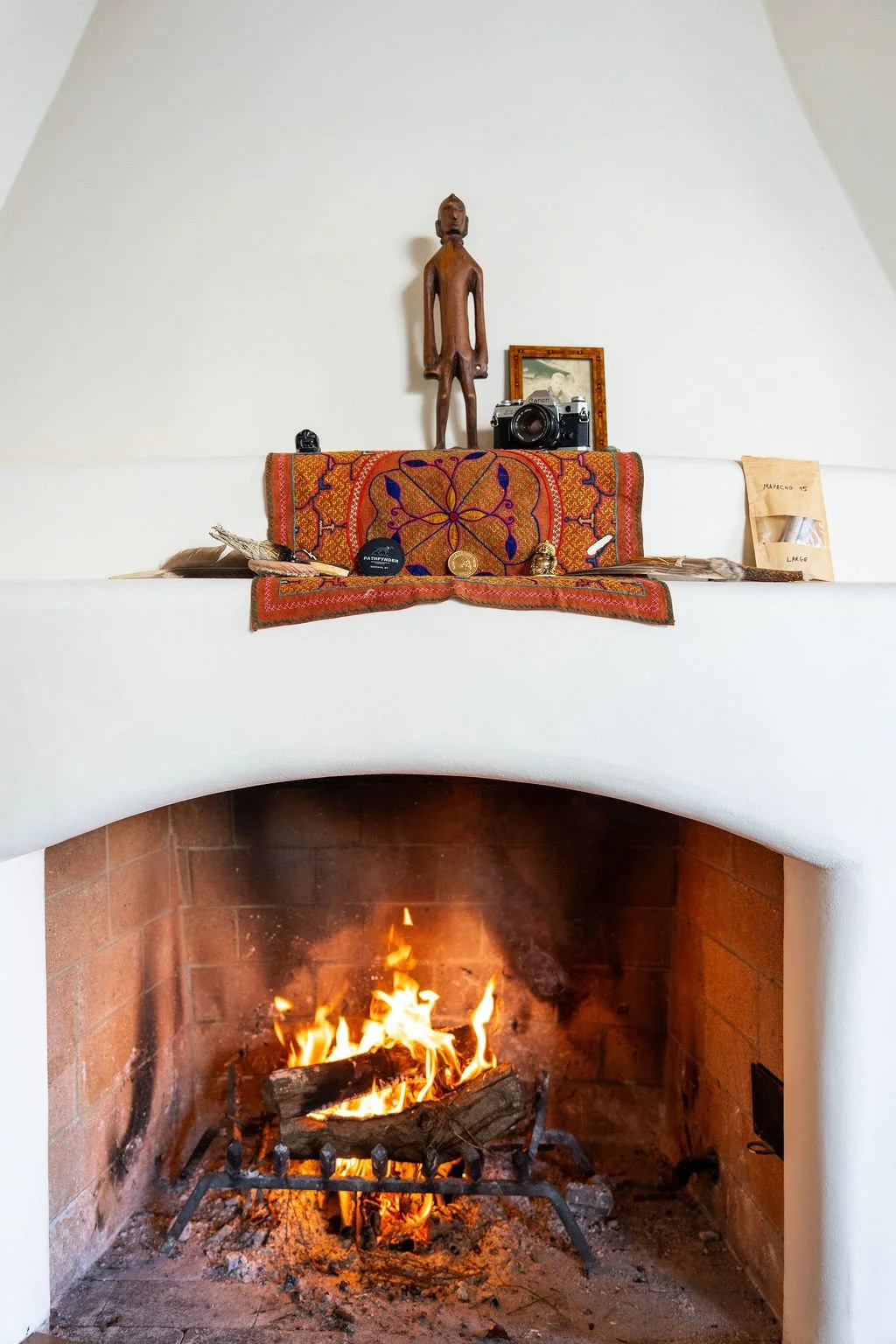 A cozy fireplace with a burning fire, decorated mantle with a colorful embroidered cloth, wooden statue, camera, small framed picture, and various small objects and packages.