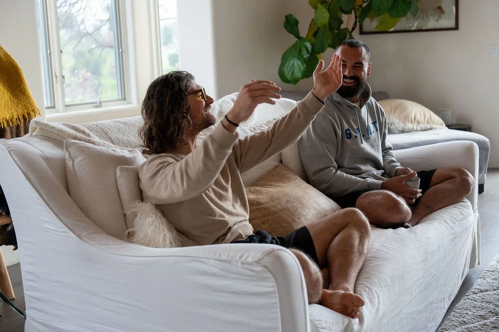 Two men sitting on a white couch in a living room, smiling and high-fiving each other.