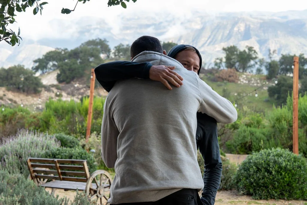 Two people hugging outdoors in a garden, with mountains and trees in the background.