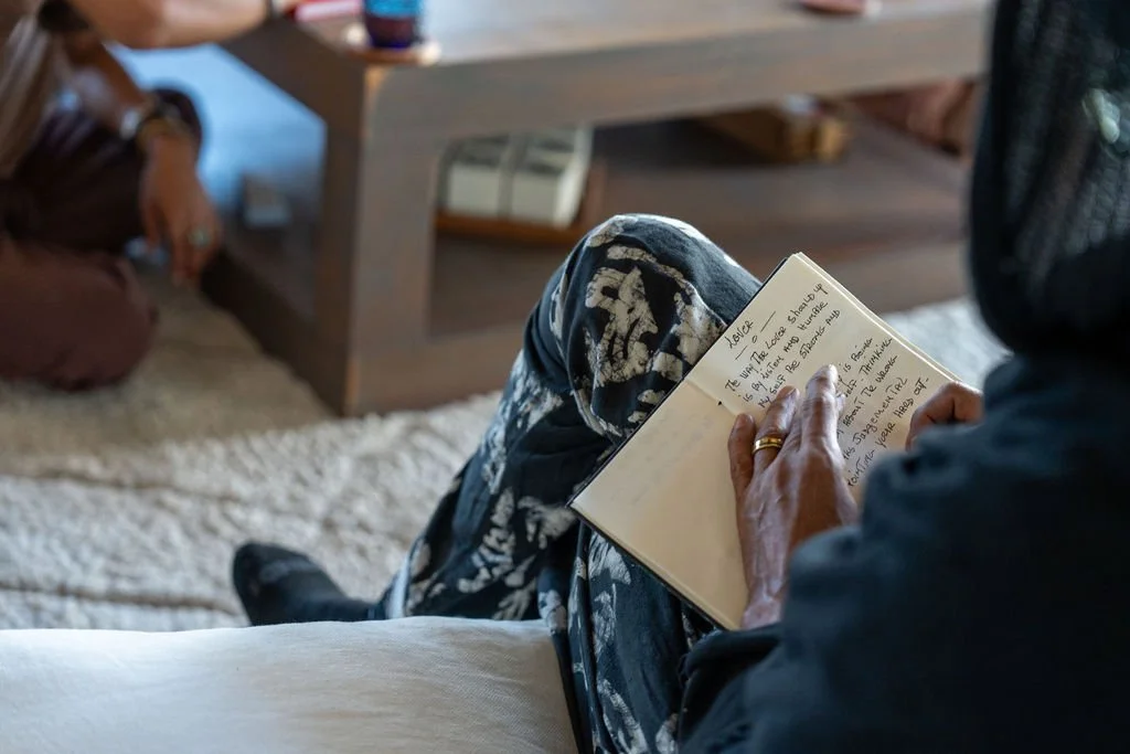 Person sitting on a couch writing in a small notebook, with another person partially visible sitting nearby and a coffee table in the background.