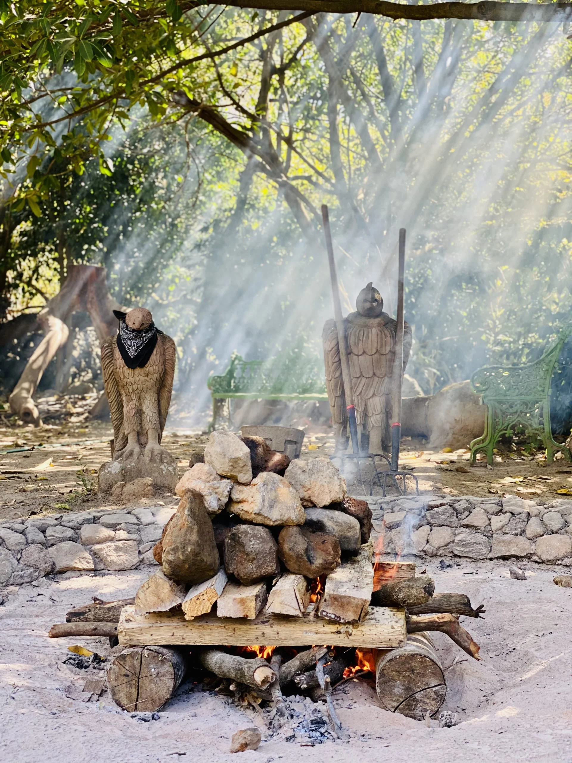 A campfire made of wood logs, with rocks surrounding it, burning outdoors in a natural setting. Two carved wooden statues with carved feathers are positioned behind the fire, with sunlight streaming through trees in the background.