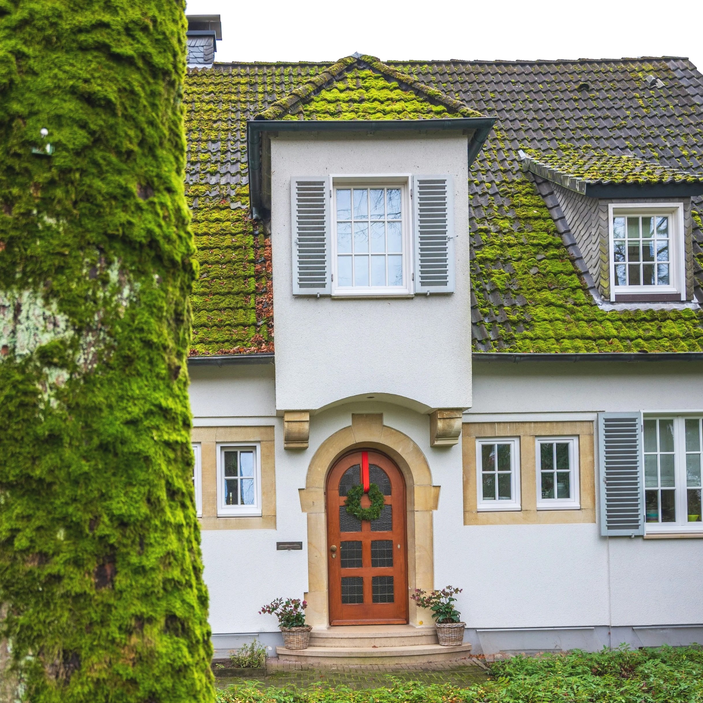 Front view of a house with a moss-covered roof, a white balcony, and a wooden front door decorated with a wreath, flanked by potted plants and surrounded by greenery.
