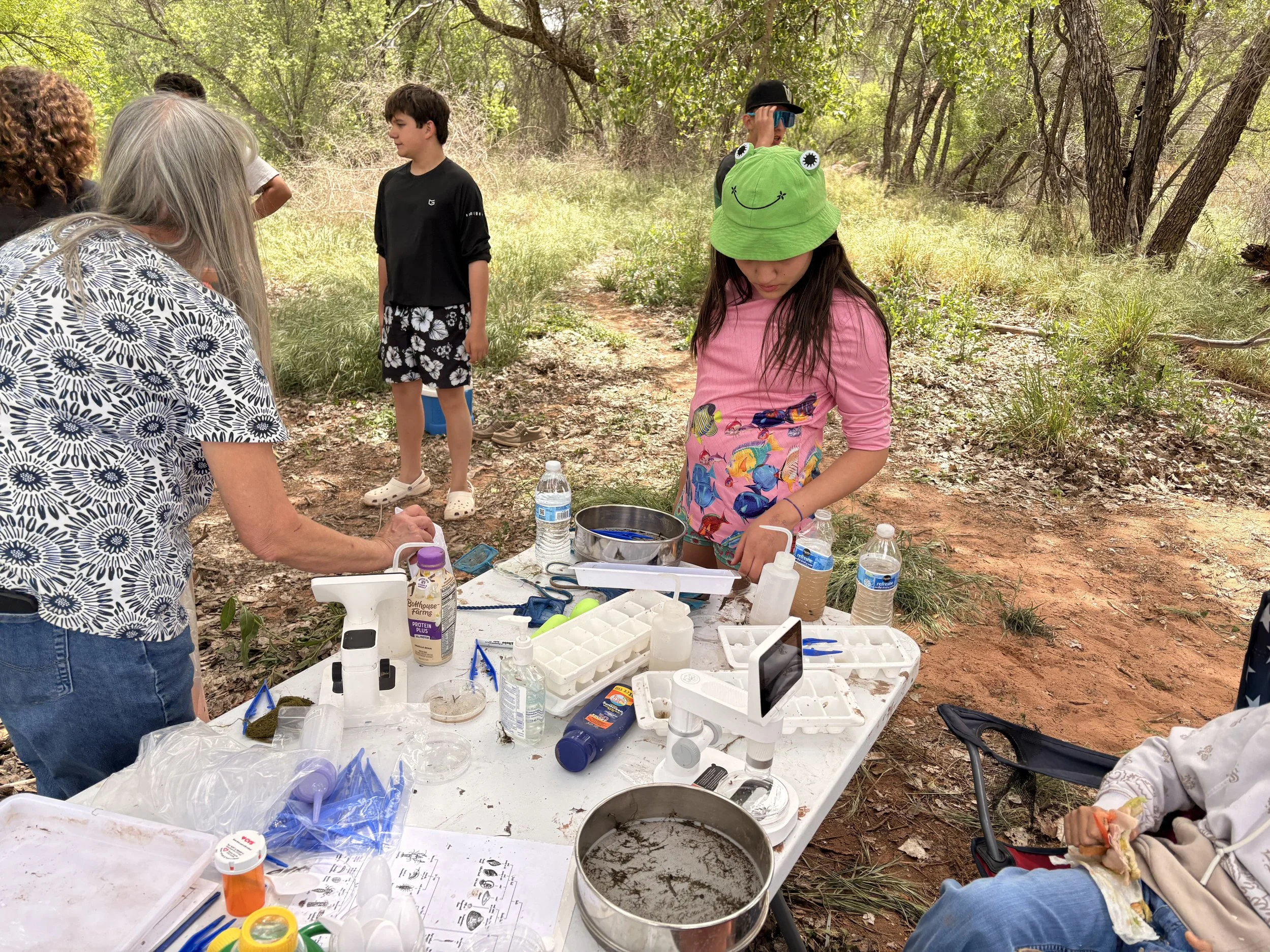 A group of children and adults participating in an outdoor science activity in a wooded area with trees and grass. They are gathered around a table with scientific equipment, bottles of water, and supplies, engaged in hands-on experiments.