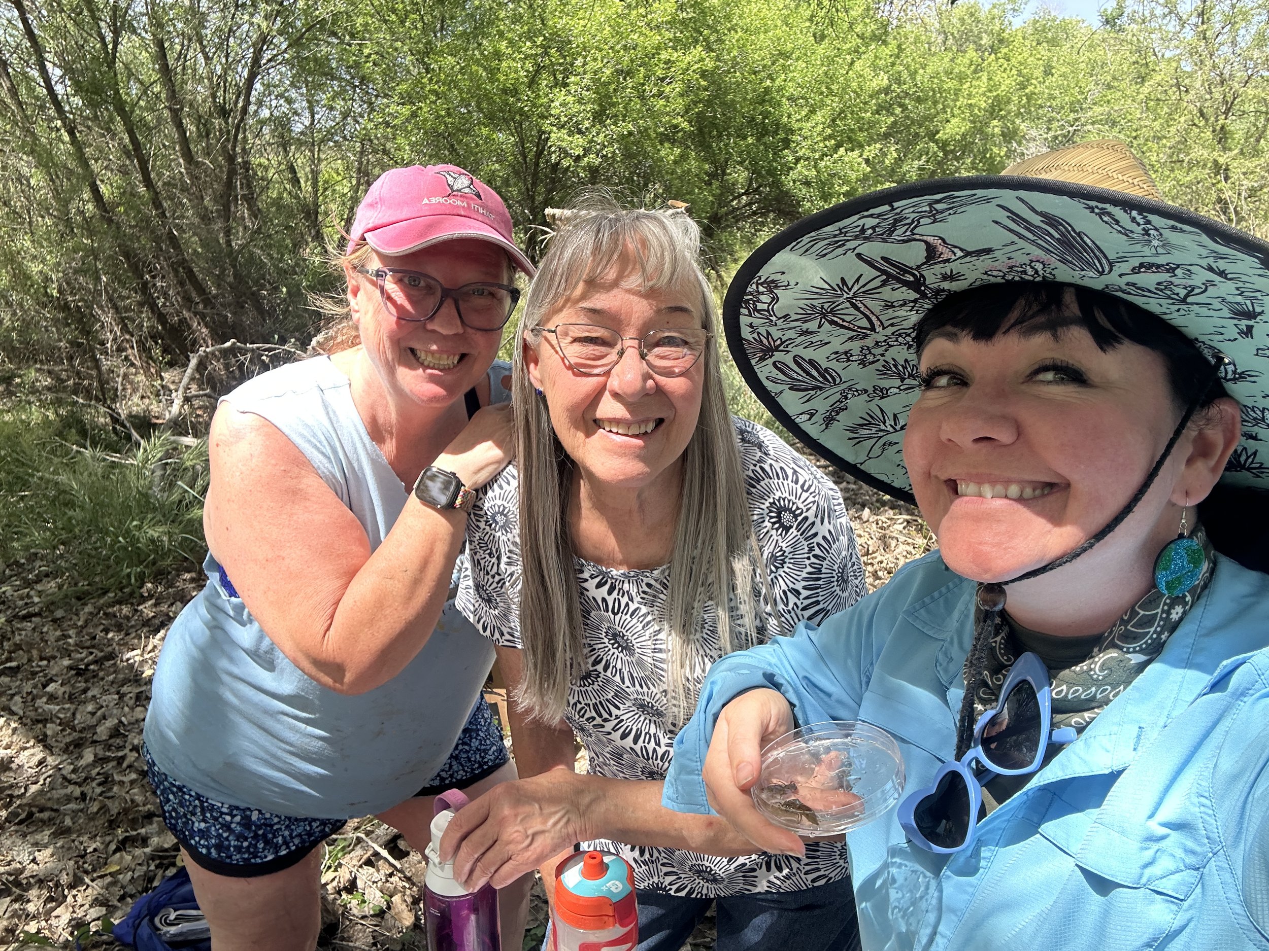 Three women outdoors in a wooded area smiling at the camera. One woman is wearing a wide-brimmed hat with a nature pattern, glasses, and a blue jacket. The second woman has glasses, gray hair, and a white shirt with a black floral pattern. The third woman is wearing a pink cap, glasses, and a sleeveless top.