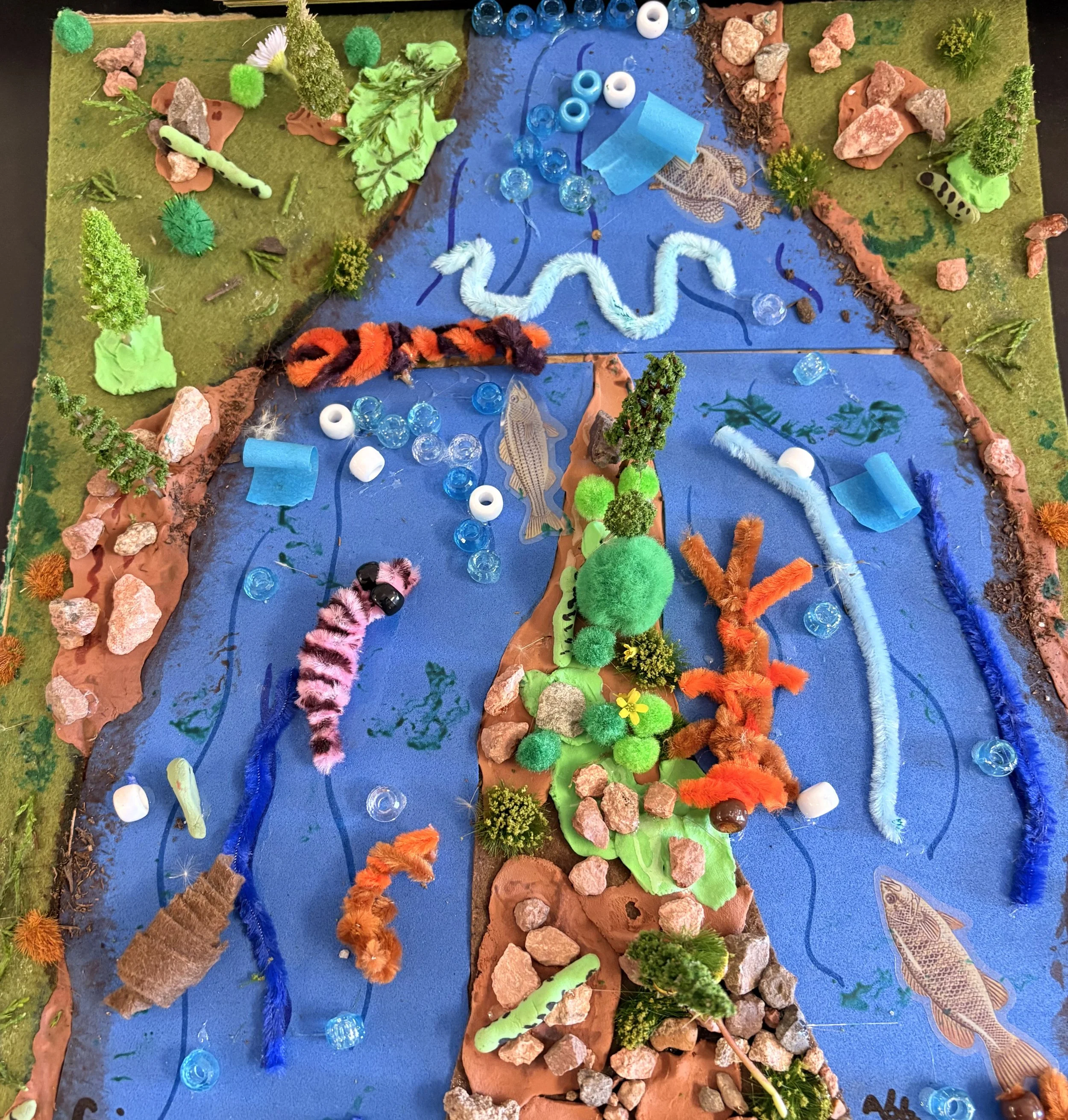 A handmade diorama of a river scene created with craft materials. The scene includes blue paper or fabric representing water, surrounded by rocks, greenery, and trees made from various craft supplies. There are small pom-poms, pipe cleaners, and beads used for decorations and details, with some animals or fish drawn or cut out and placed within the scene.