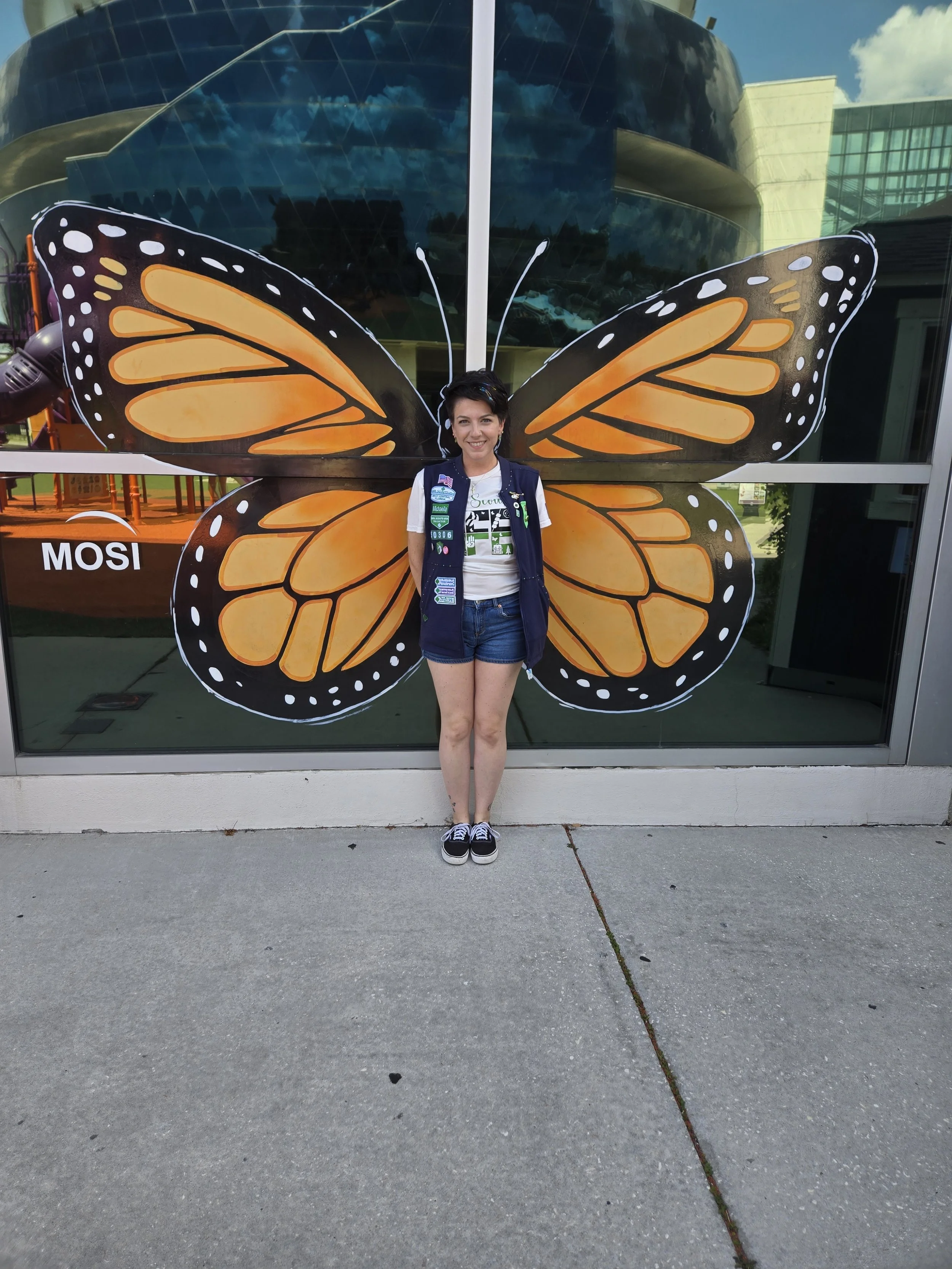 A woman standing in front of a glass building with a large butterfly-shaped decoration behind her. She is smiling and wearing a white T-shirt, denim shorts, and a navy blue vest with various patches.