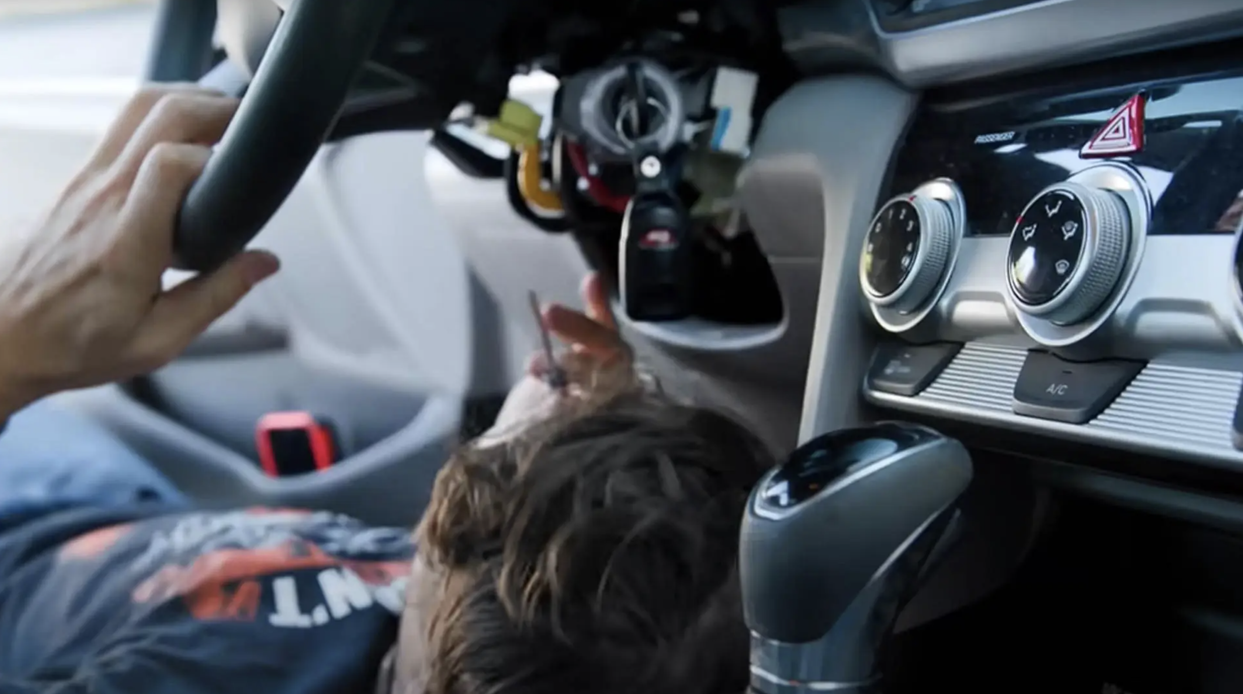 Inside a car, a person's hand is on the steering wheel, and another person's head is visible, with car controls and dashboard visible in the background.
