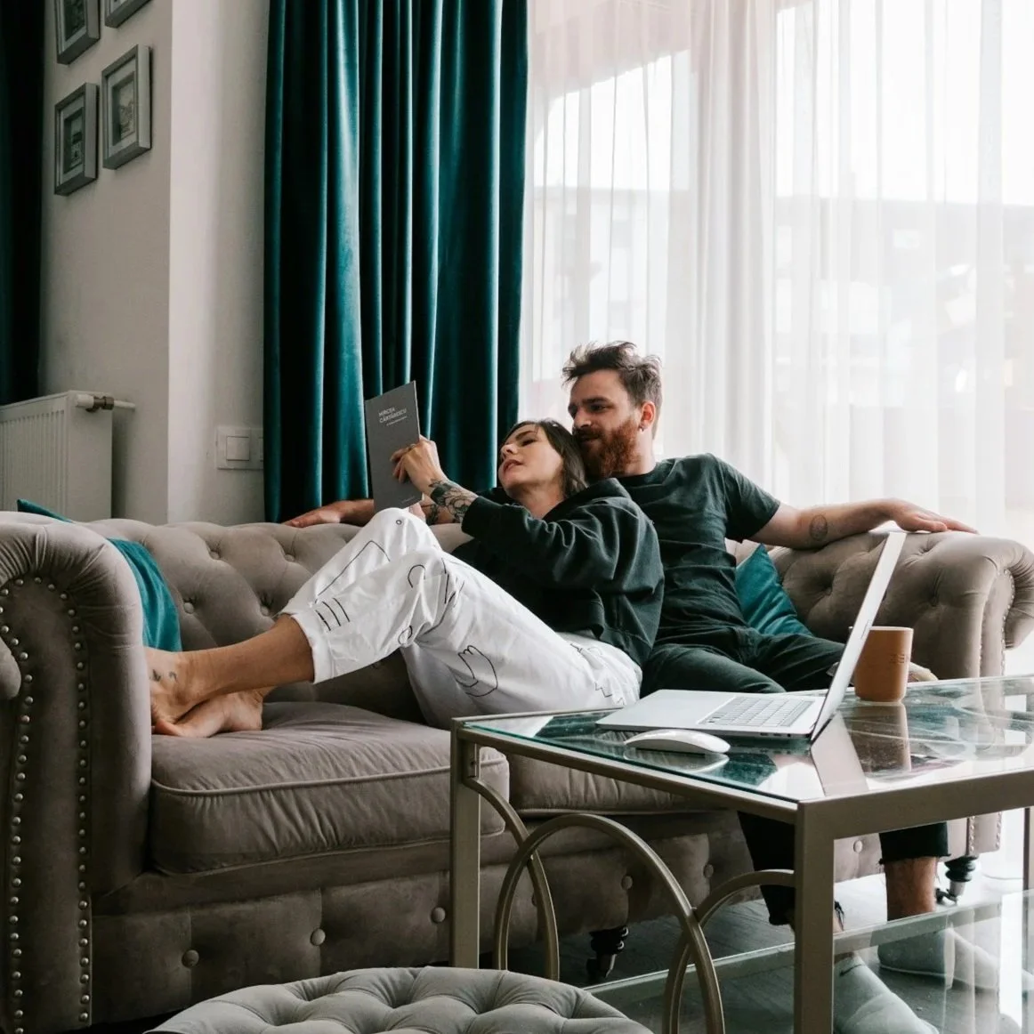 A man and woman sitting on a beige couch, looking at a book together, with a laptop on a glass coffee table in front of them, in a bright living room with white curtains and teal drapes.