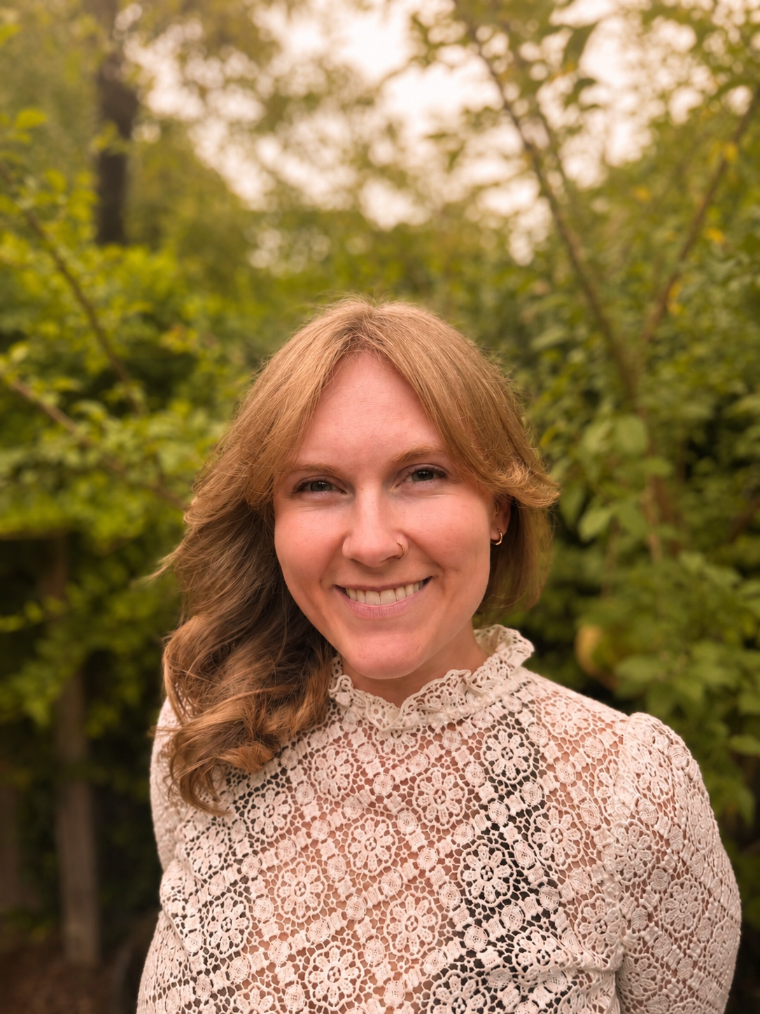 Portrait of a young woman with light skin and reddish-brown hair, smiling, wearing a cream-colored lace top, outdoors in a green, tree-filled environment.