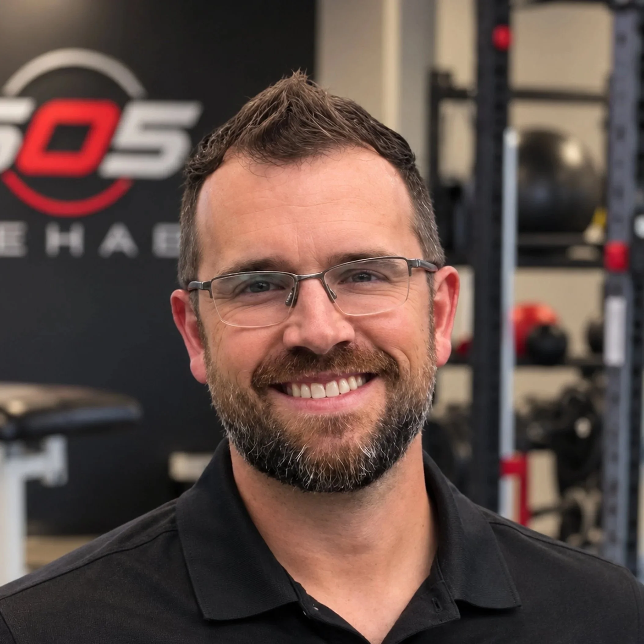 Close-up of a man with glasses and a beard, smiling, in a gym or workout area.