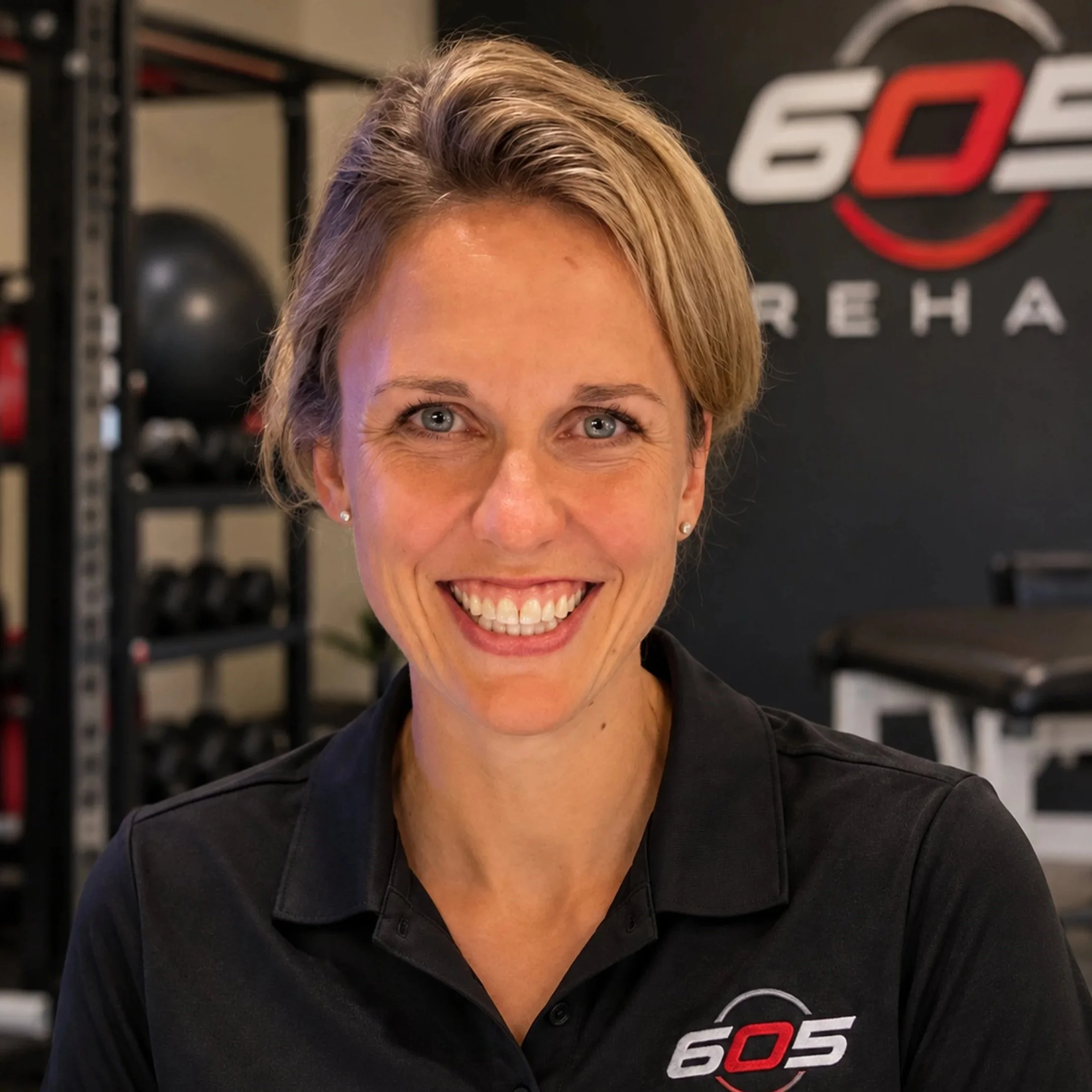 Close-up of a woman's face with blonde hair and blue eyes, smiling. She is in a fitness center or gym, with fitness equipment and a logo reading '605' in the background.
