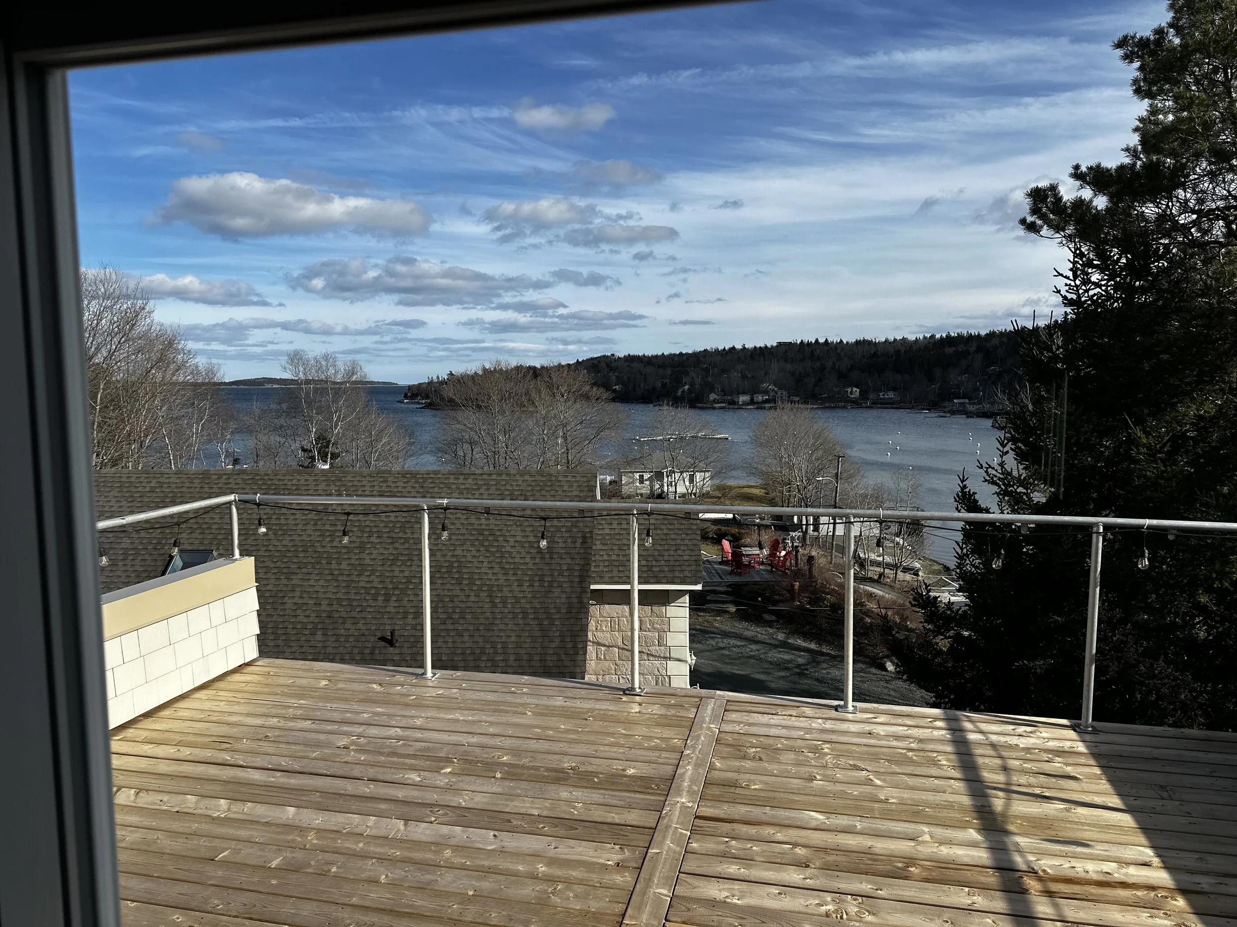 View through window of a lake or river with trees and a small island, blue sky with clouds, and the roofs of nearby houses, taken from a balcony or deck with wooden flooring and metal railing.