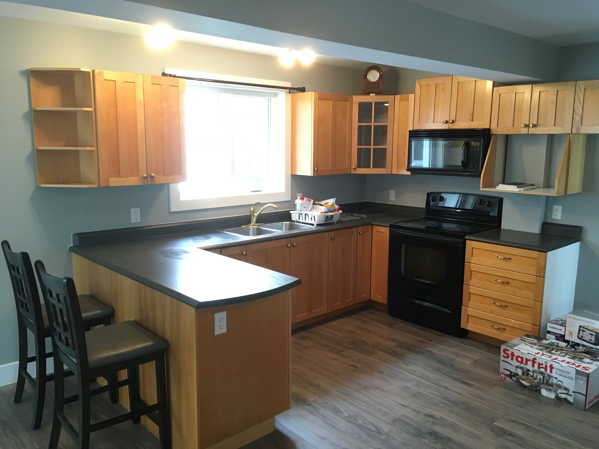 Kitchen with wooden cabinets, black appliances, and a countertop island with four black chairs. There is a window above the sink, and various boxes and items on the floor and counter.