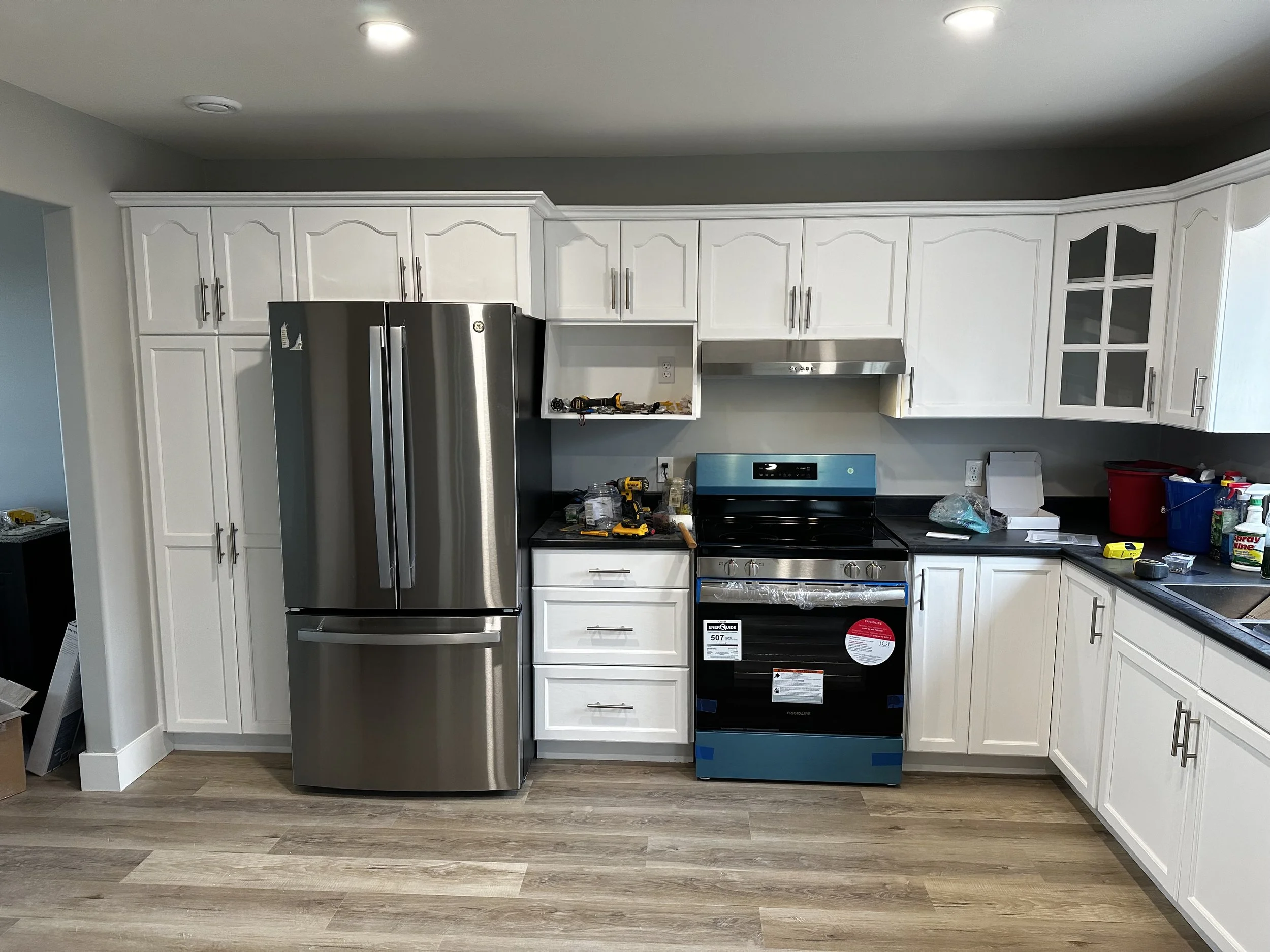 Kitchen with white cabinets, stainless steel refrigerator to the left, black stove with oven, various tools and items on the counter, and partially installed range hood.