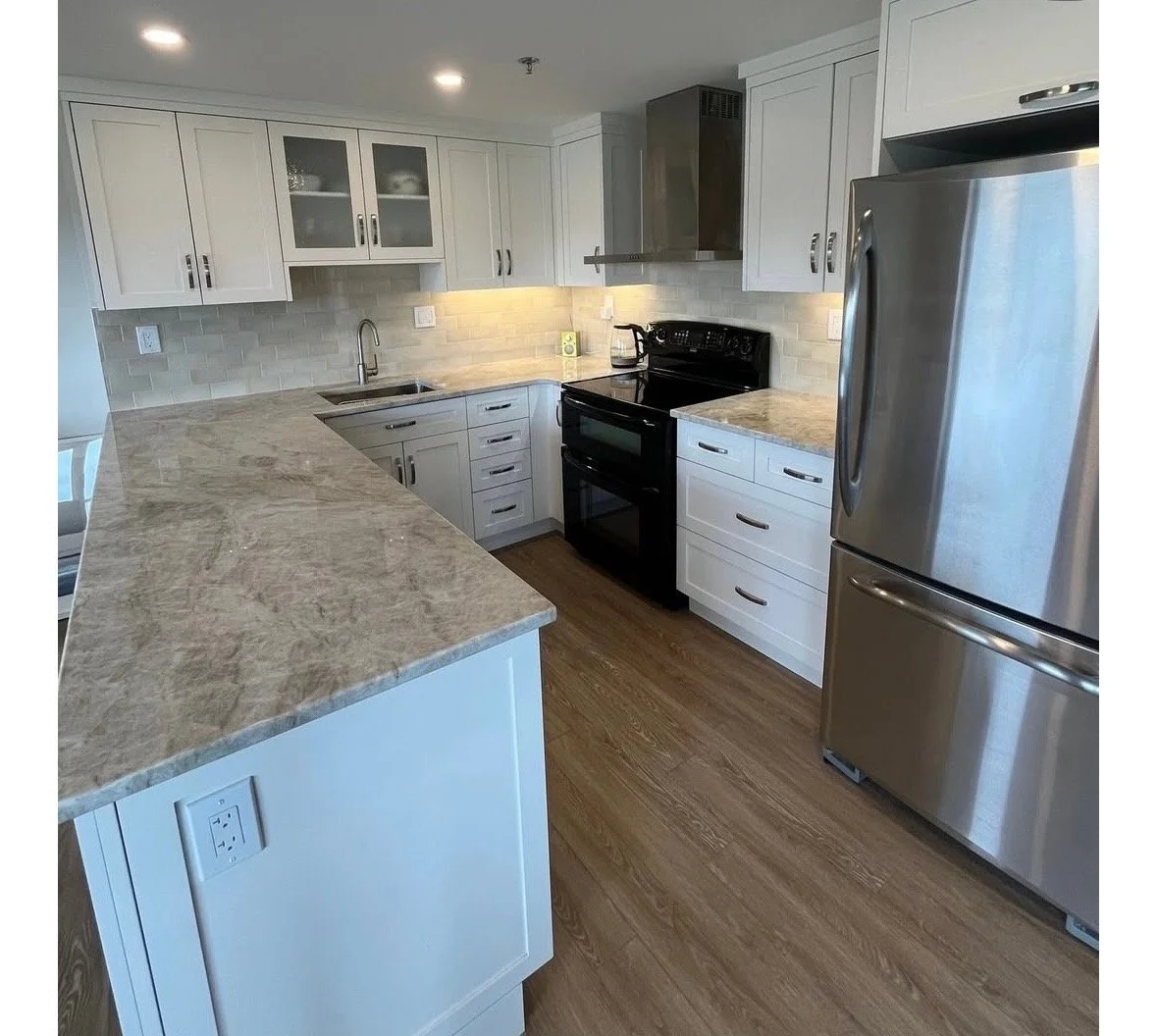 Modern kitchen with white cabinets, beige granite countertops, stainless steel appliances including a refrigerator and oven, tan brick backsplash, and wooden flooring.
