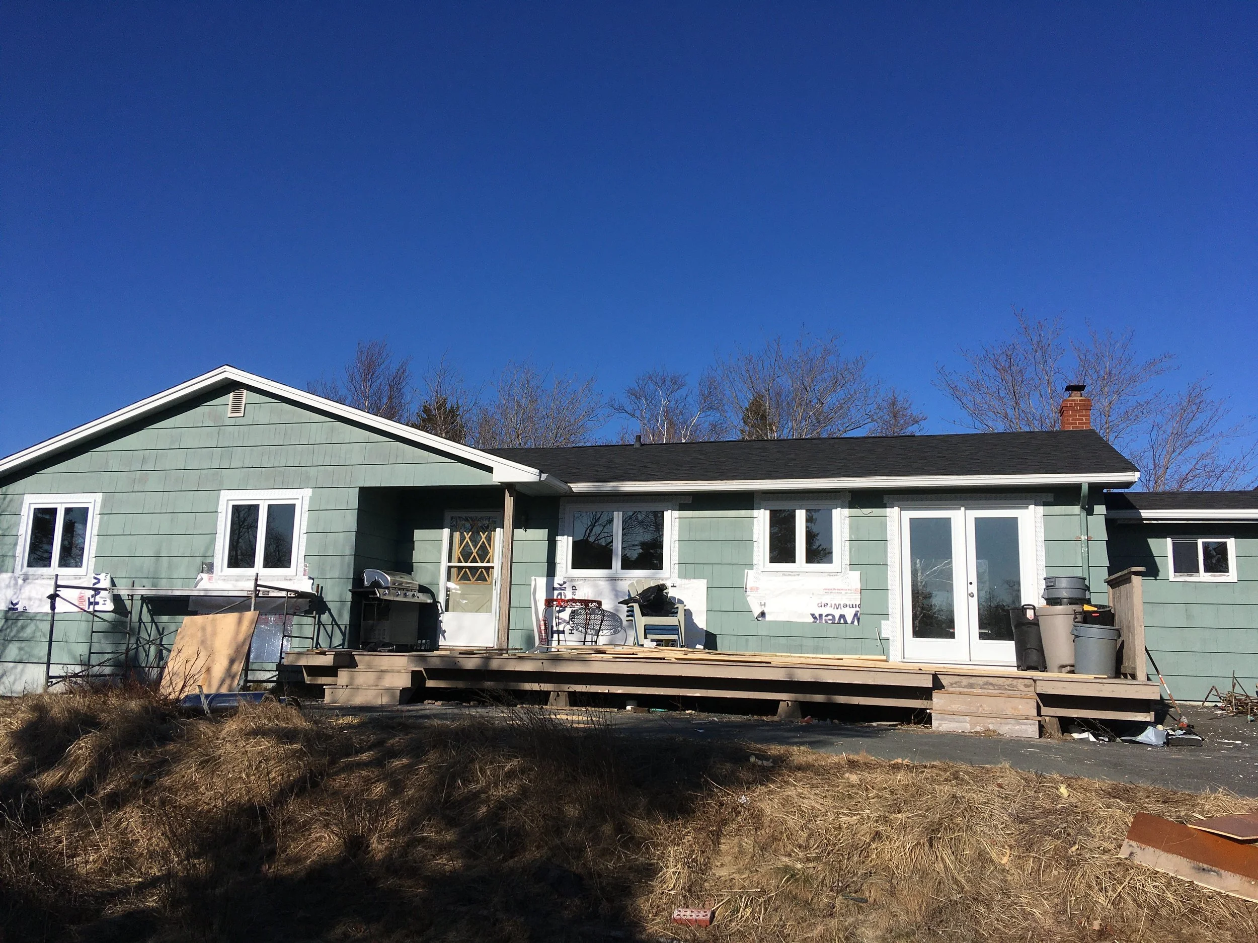 A house under construction with green exterior siding, multiple white-framed windows, and a deck in progress. Construction materials and tools are scattered in the yard, and there are leafless trees and a bright blue sky in the background.