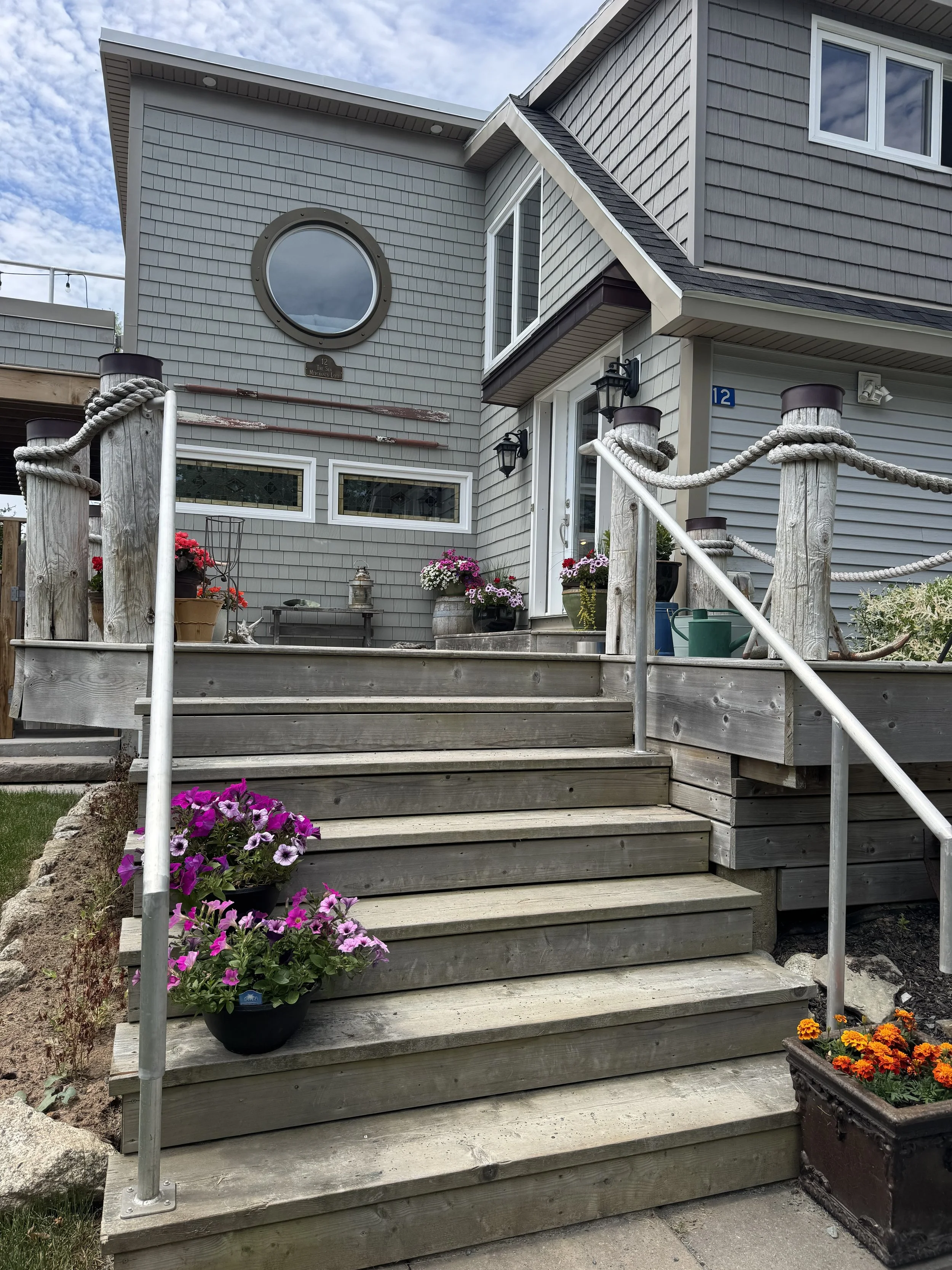Front porch stairs with potted flowers including pink, purple, and orange blooms, leading up to a house with gray shingle siding, black outdoor lanterns, and a blue house number 12. There is a round window and rectangular windows on the house, with a
