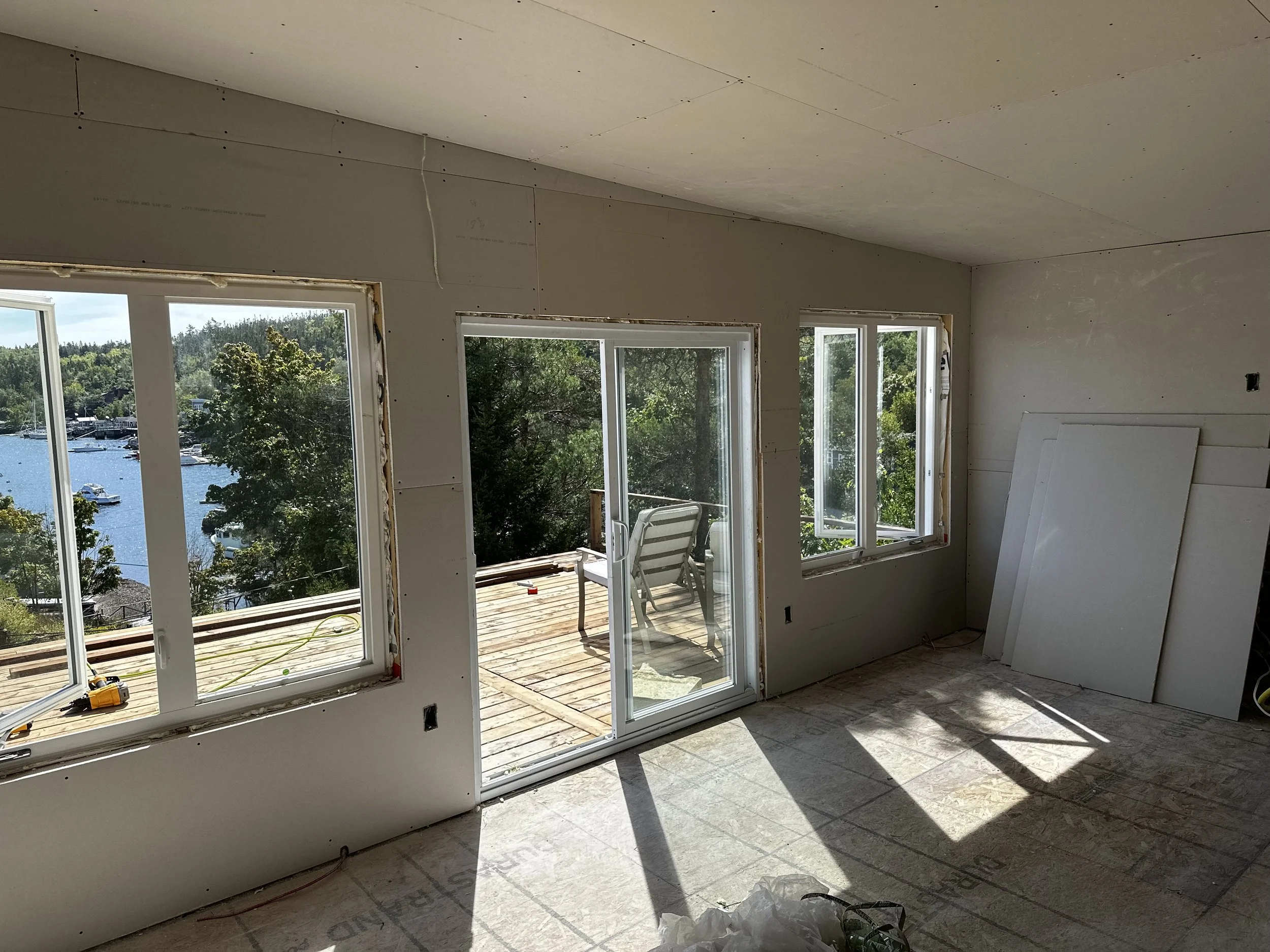 An unfinished room with large windows and sliding glass doors overlooking a body of water with boats, and trees. The room has drywall installed on the walls and ceiling, and construction tools and materials are present.