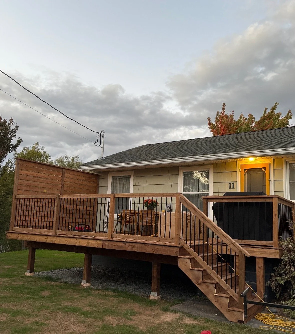 New wooden deck with stairs attached to the back of a house with a light on above the door, a table with chairs, and a barbecue grill, during cloudy weather.