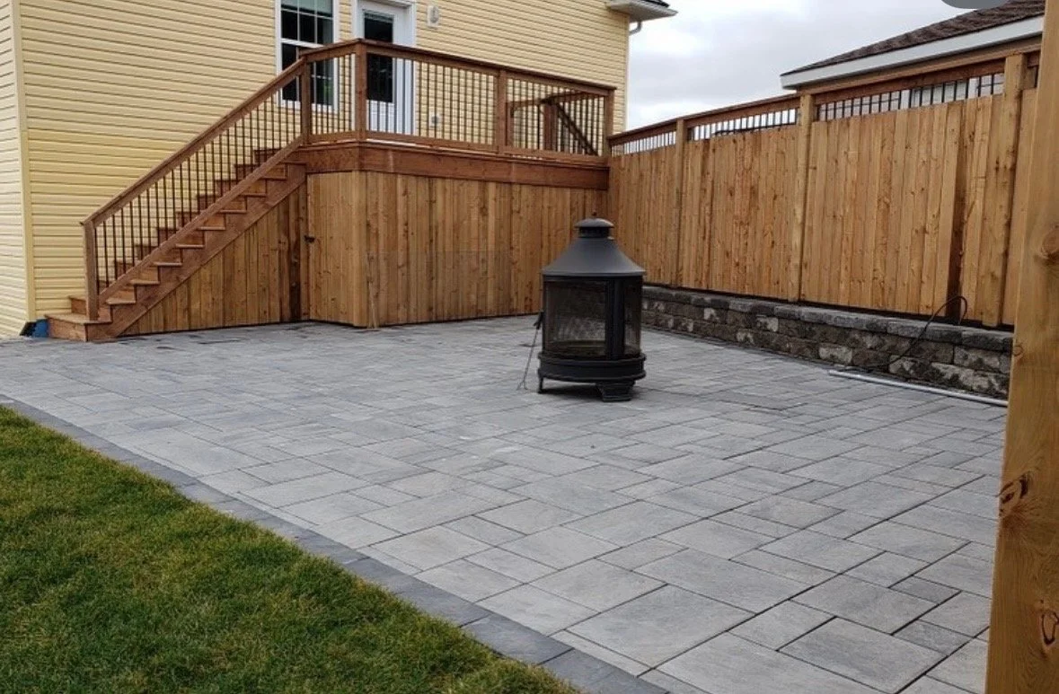 Backyard patio with a stone tile surface, a wooden deck with stairs, a wooden privacy fence, a small patch of grass, and a black outdoor heater or fire pit near the center.