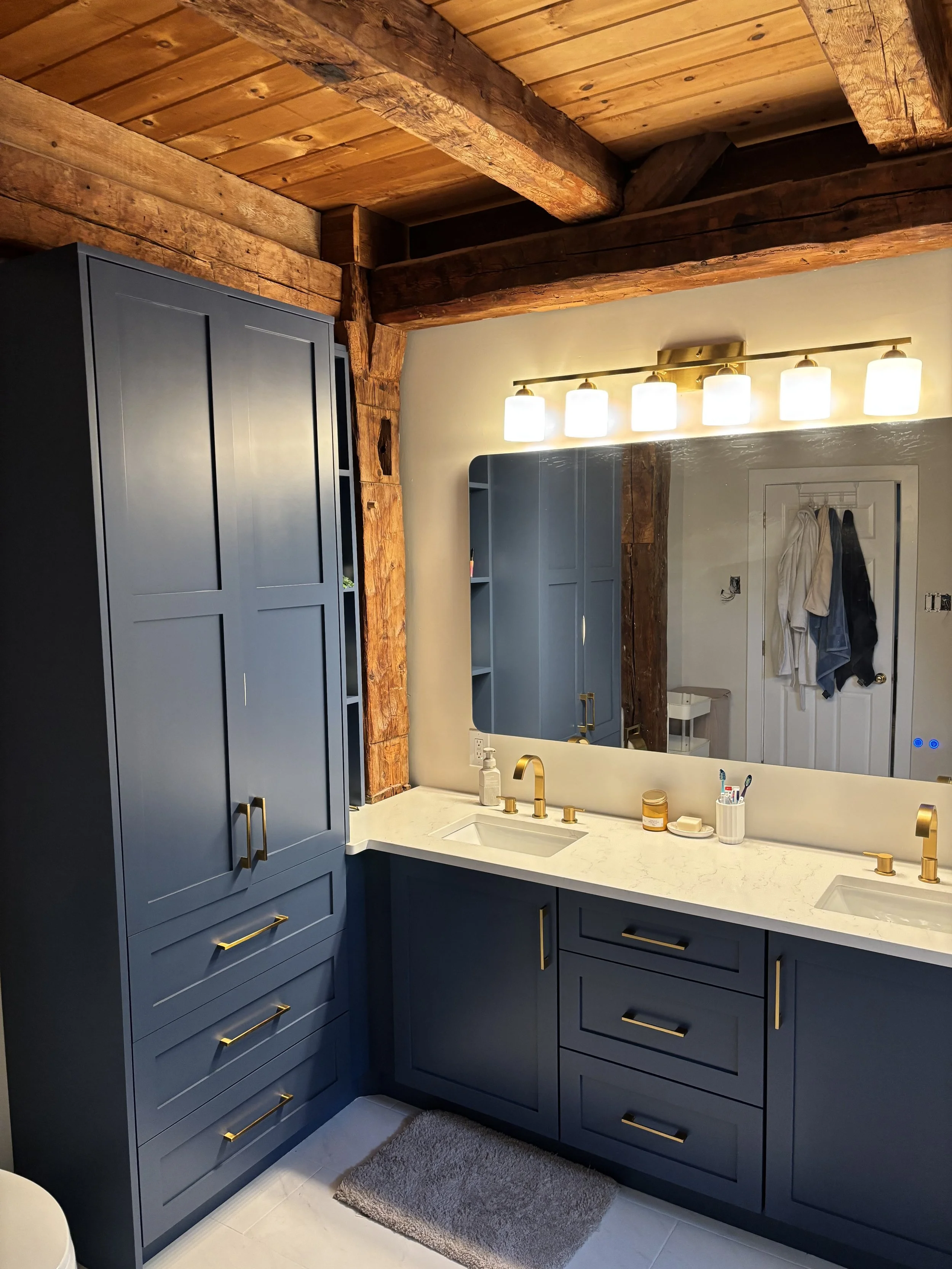 A modern bathroom with blue cabinets, a white marble countertop, gold fixtures, and a large mirror. There are wooden beams on the ceiling and a row of four bright vanity lights. The bathroom has a small rug, and items like toothbrushes, soap, and lot