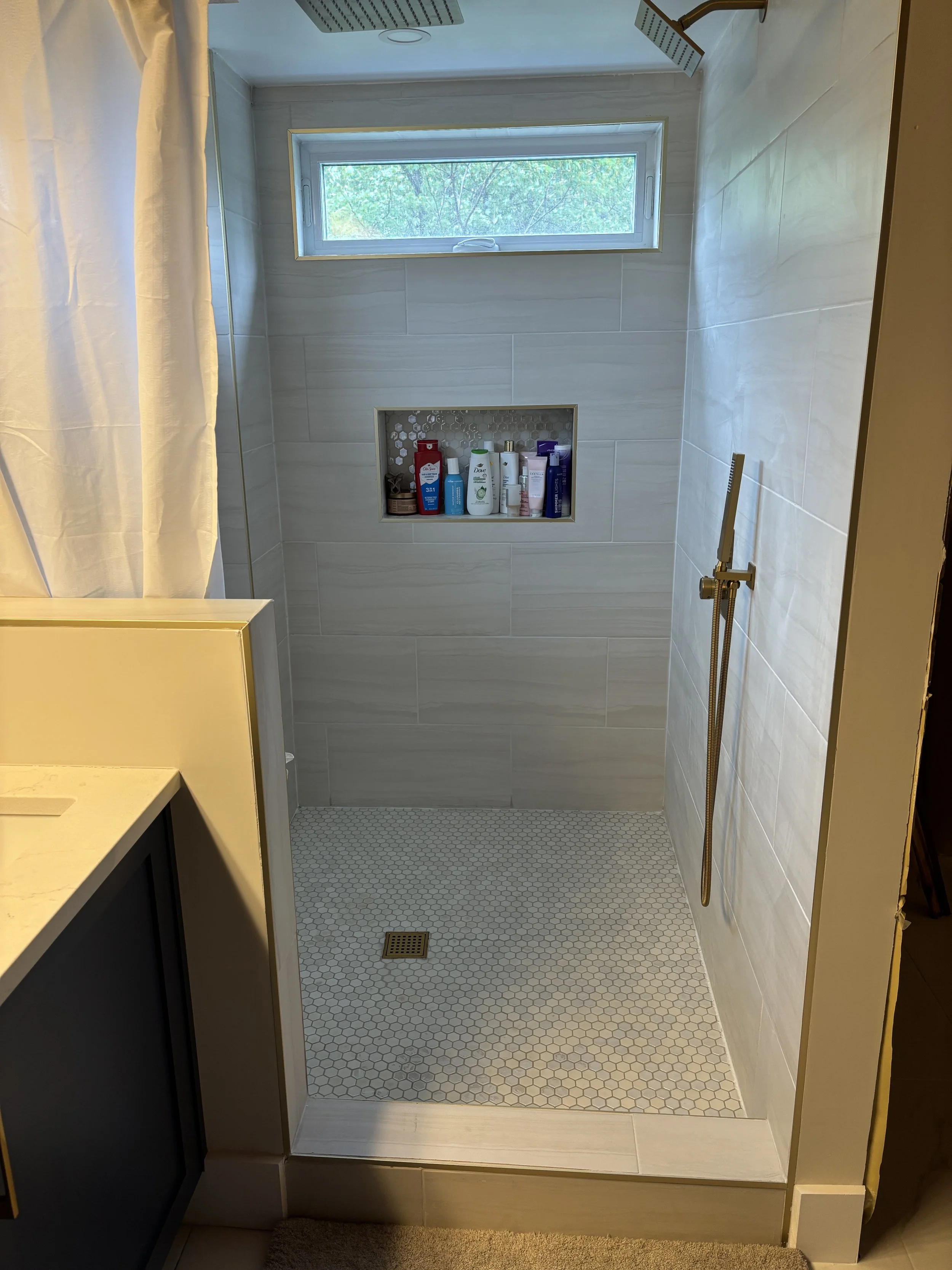 A walk-in shower with light gray tile walls, a small built-in shelf holding shampoo and body wash, a window showing greenery outside, and a shower head on the ceiling.