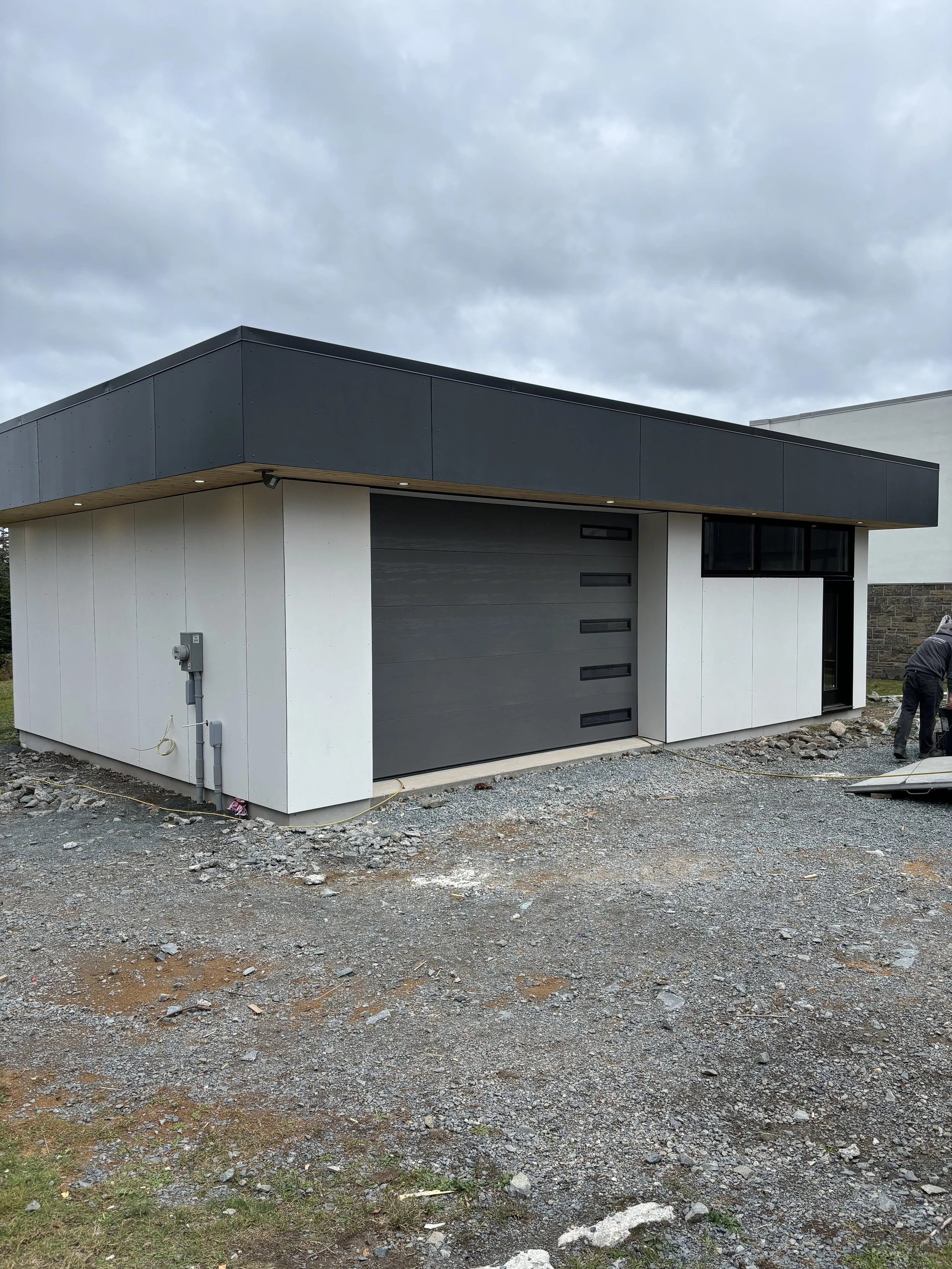 A modern house with a flat roof, black on the top and gray on the garage door, white walls, and large windows, under a cloudy sky, with construction materials and workers nearby.