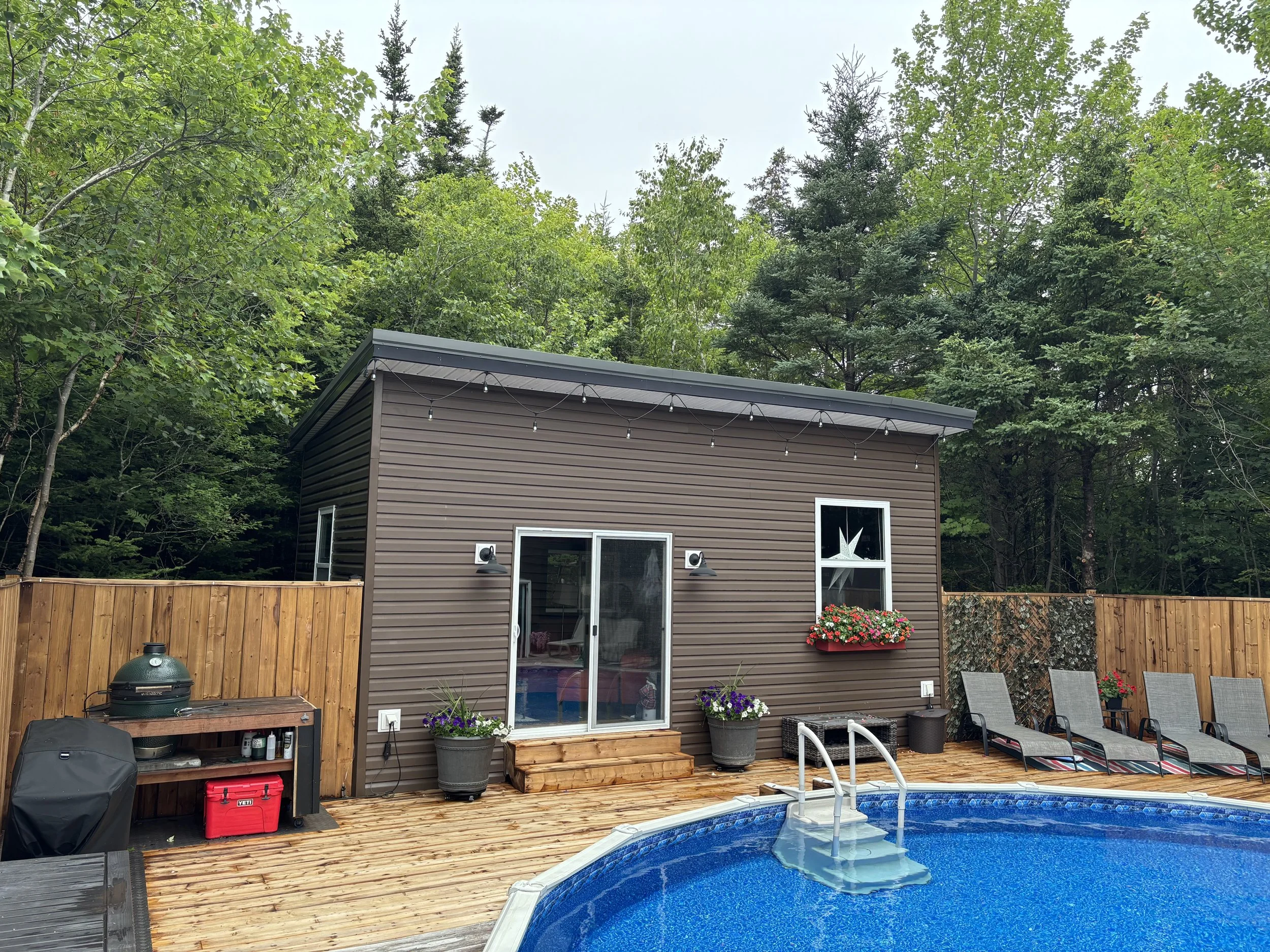 A backyard with a wooden deck, a swimming pool, and a brown house with sliding glass doors. The deck has lounge chairs, potted flowers, and a wooden step. The background features green trees and a cloudy sky.