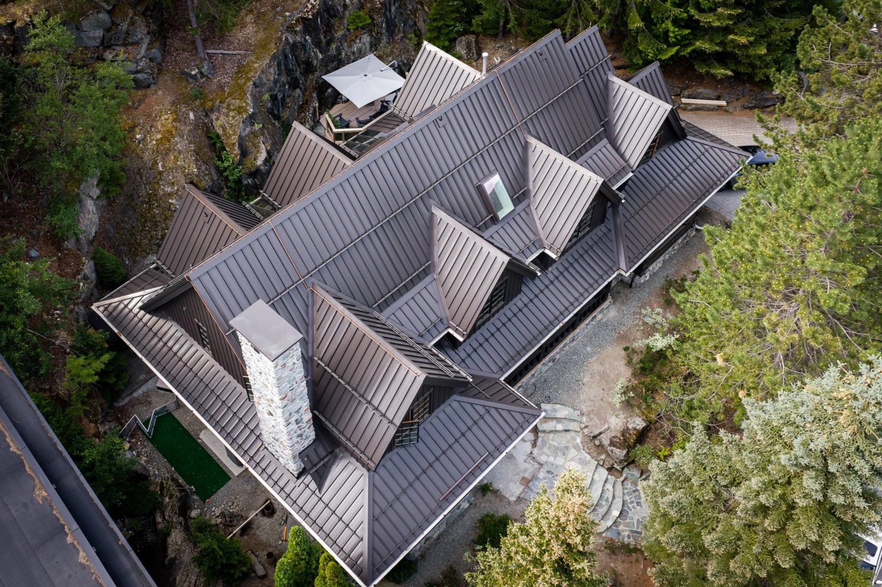 Aerial view of a custom mountain home featuring a standing seam metal roof, stone chimney, and architectural sheet metal details surrounded by forest.
